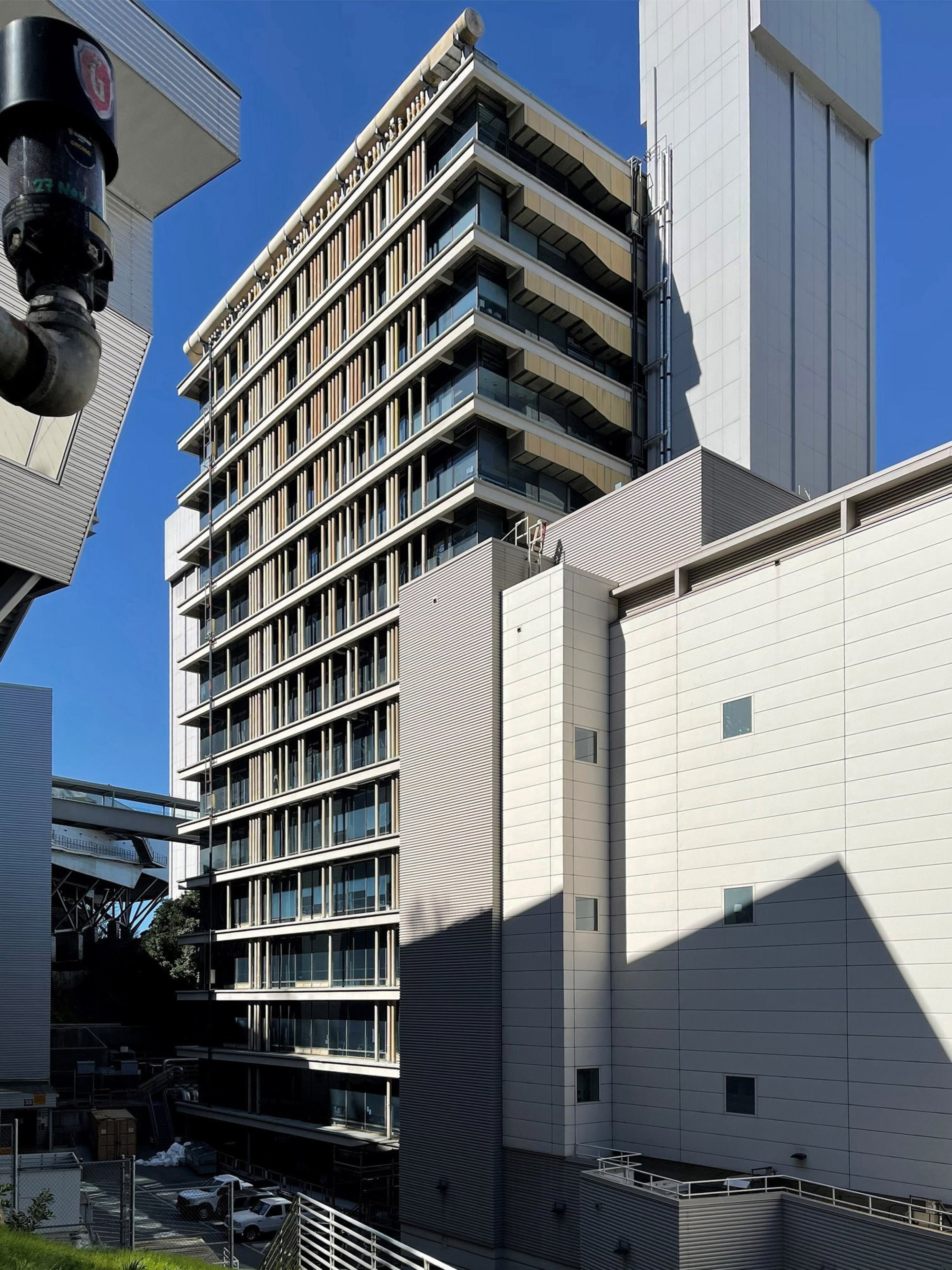 A modern, multi-story office building with large glass windows and beige accents stands against a clear blue sky. Nearby, there's another white building.