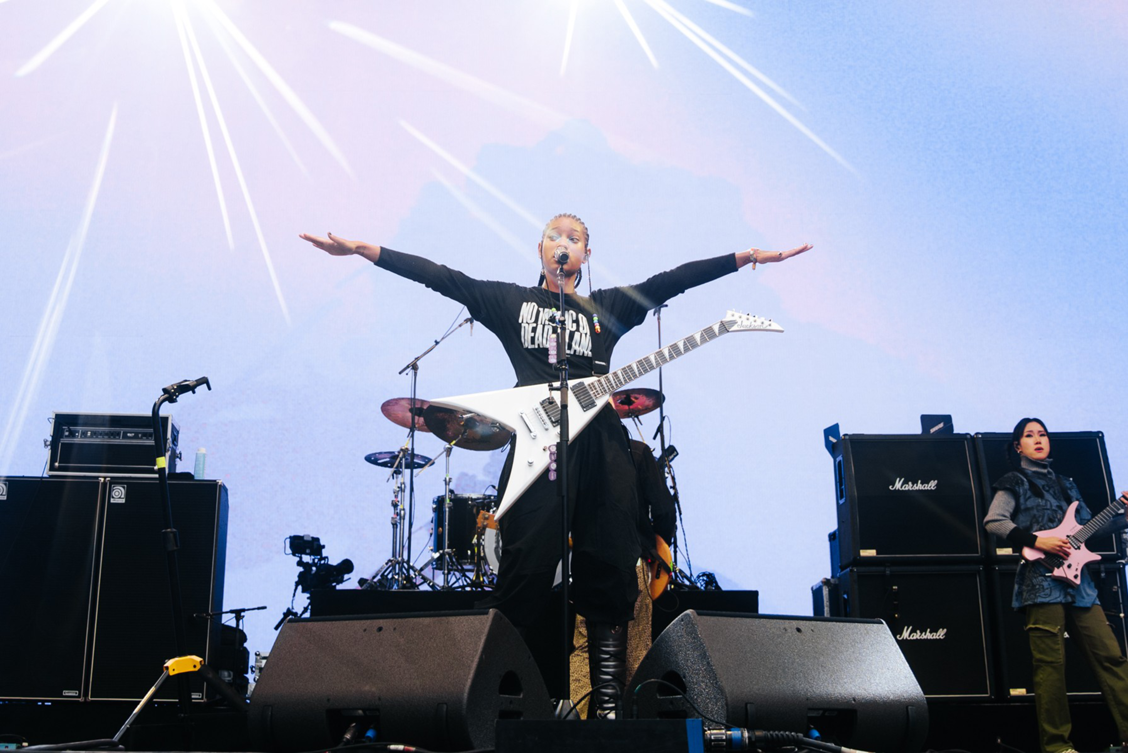 A musician stands center stage with arms spread wide, holding a white electric guitar, while another plays guitar near large Marshall amplifiers.
