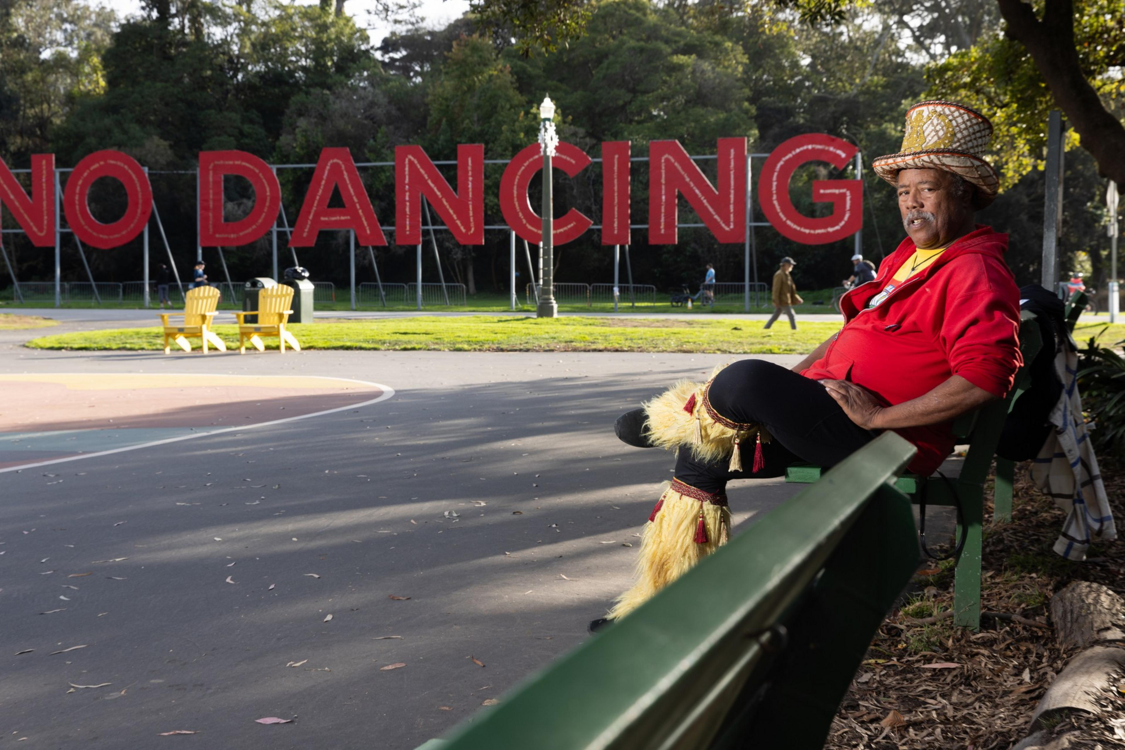 A person with a top hat and red sweater sits on a green bench with a &quot;No Dancing&quot; sign in the back.