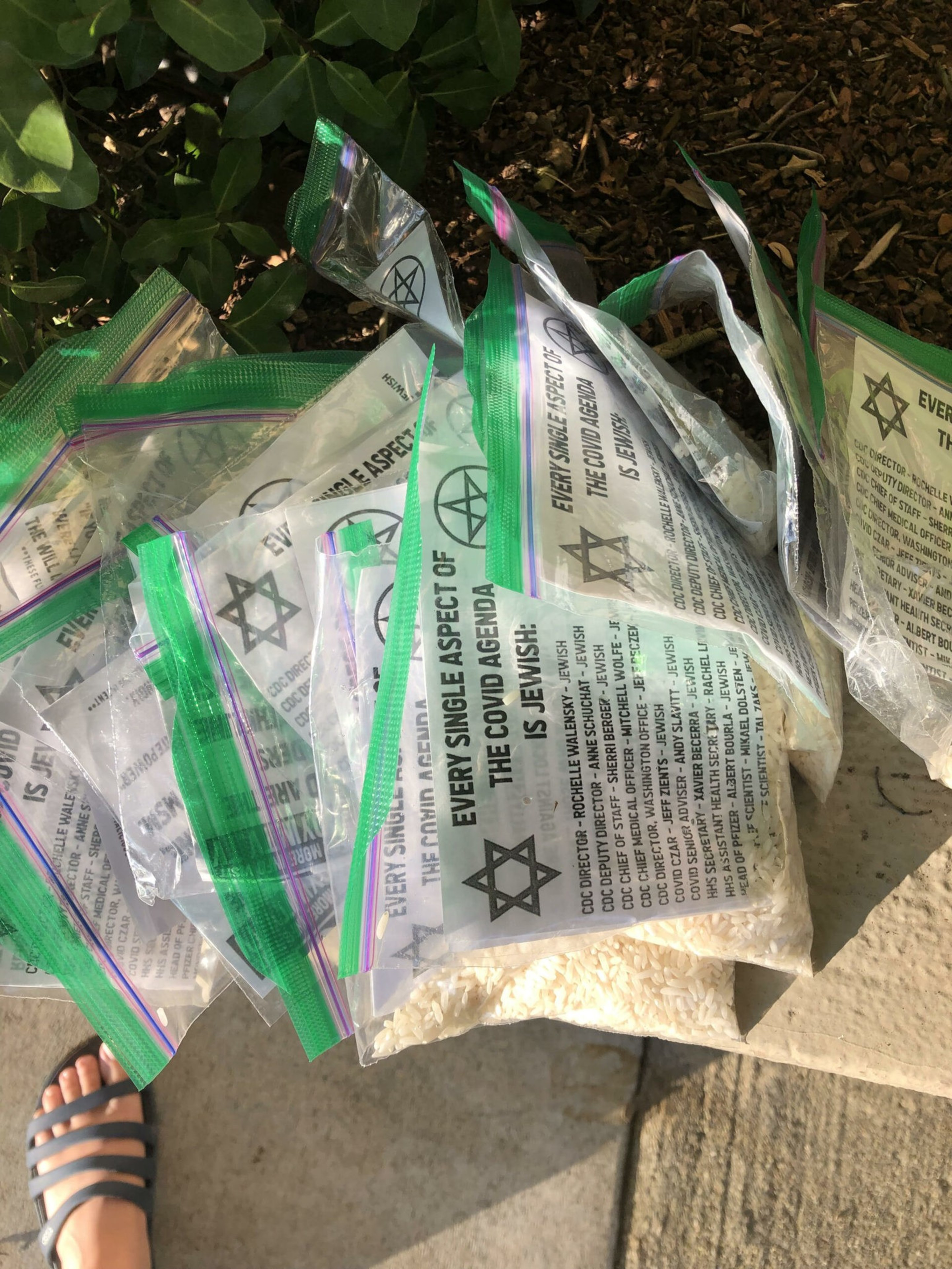 Several clear plastic bags with green zip seals contain rice and papers printed with a Star of David and text saying, "Every single aspect of the Covid agenda is Jewish."