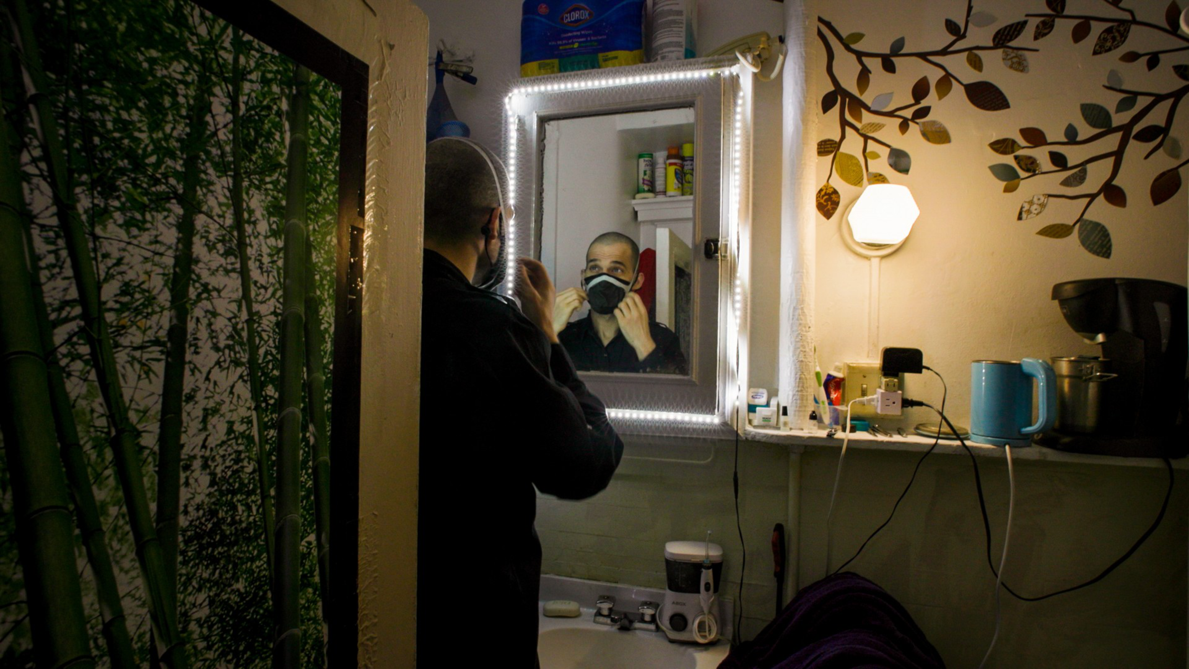 A man adjusts a black face mask while looking into a lit mirror in a dim bathroom with a bamboo wall decal and various items on a shelf.