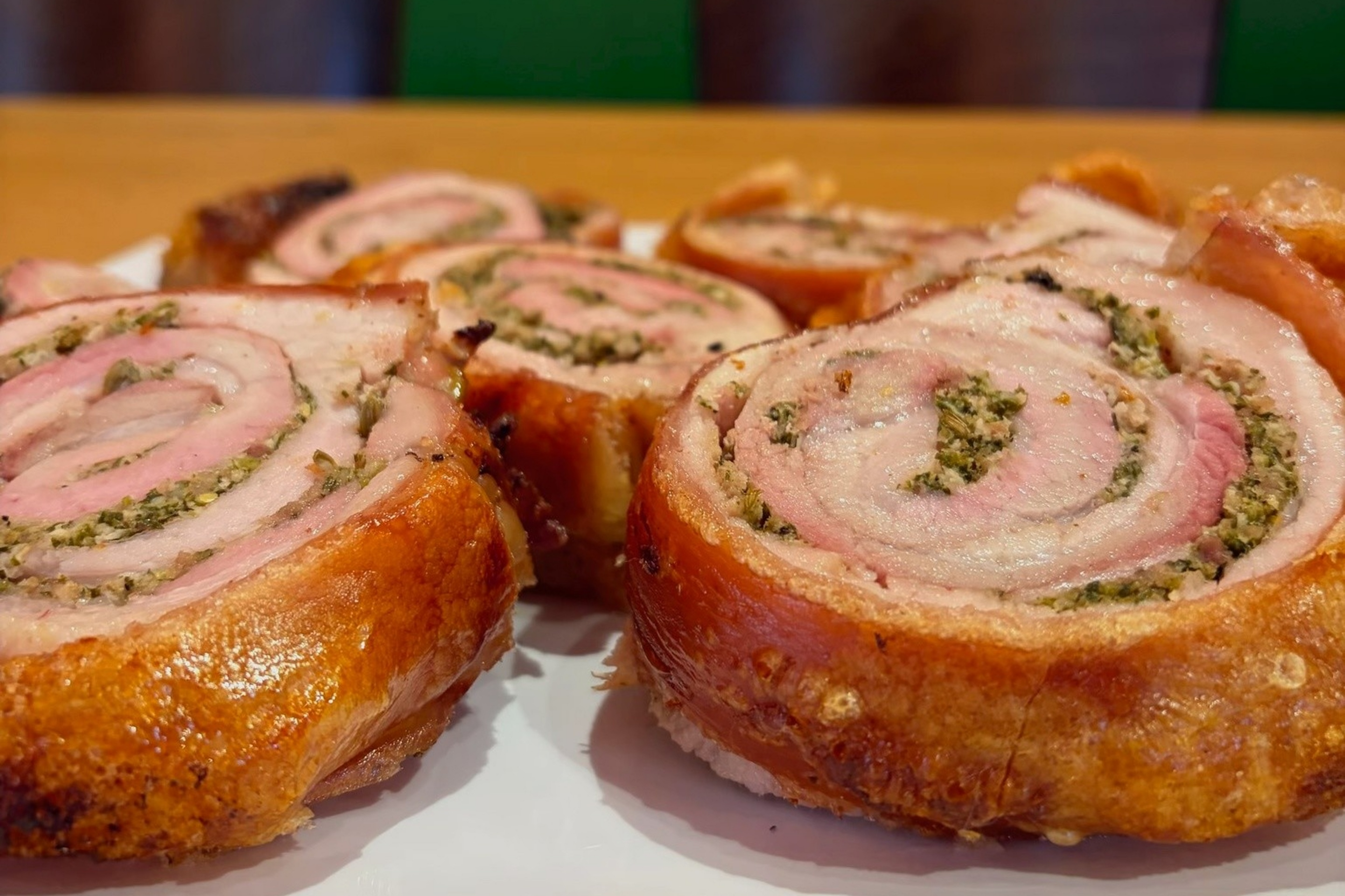 Several slices of rolled, roasted pork with herb filling are arranged on a white plate atop a wooden table, with green and peach walls in the background.