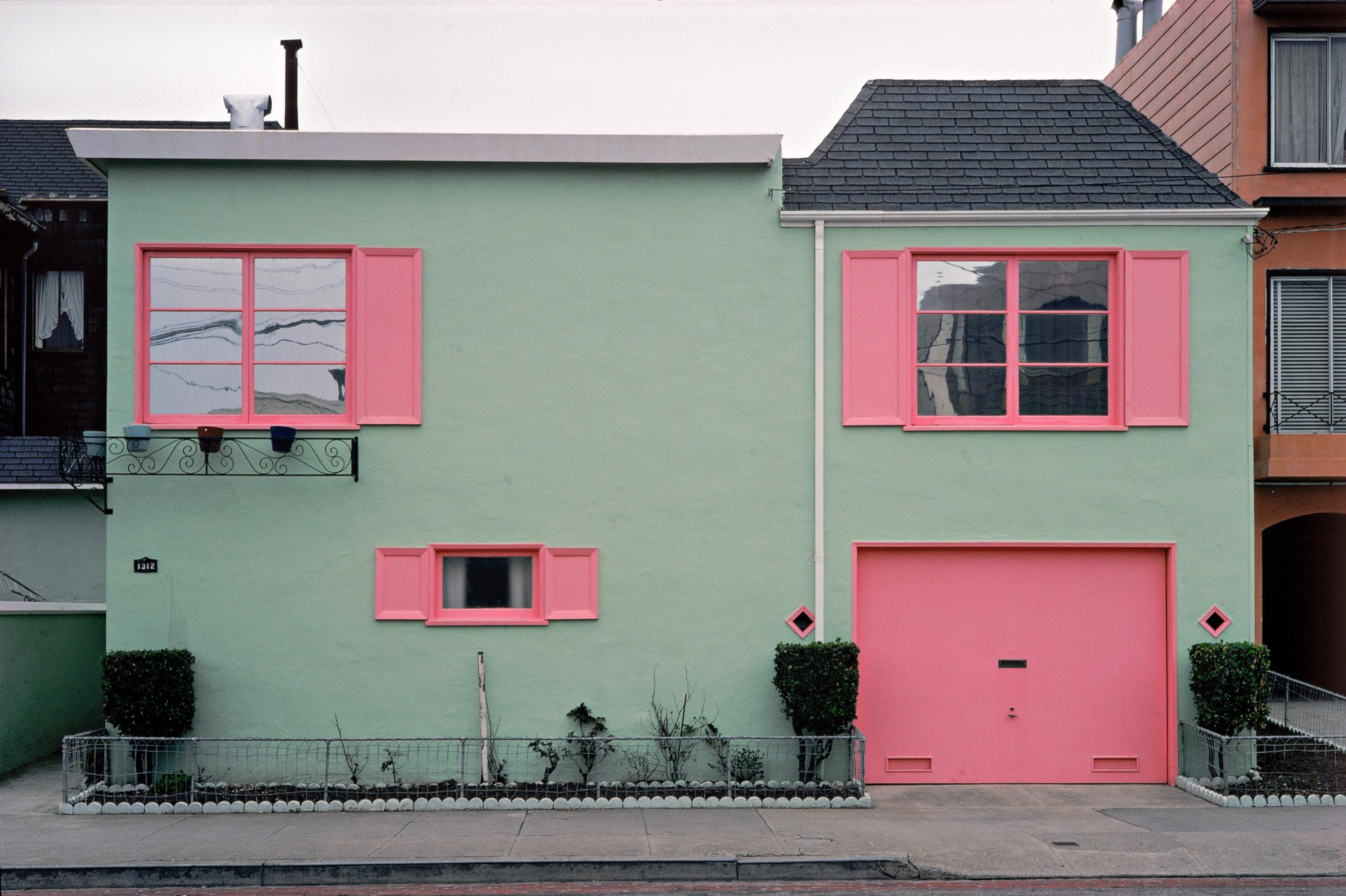 The image shows a house with pastel green walls and bright pink window frames and garage door. Small shrubs flank the entrance, and pots are on a ledge.