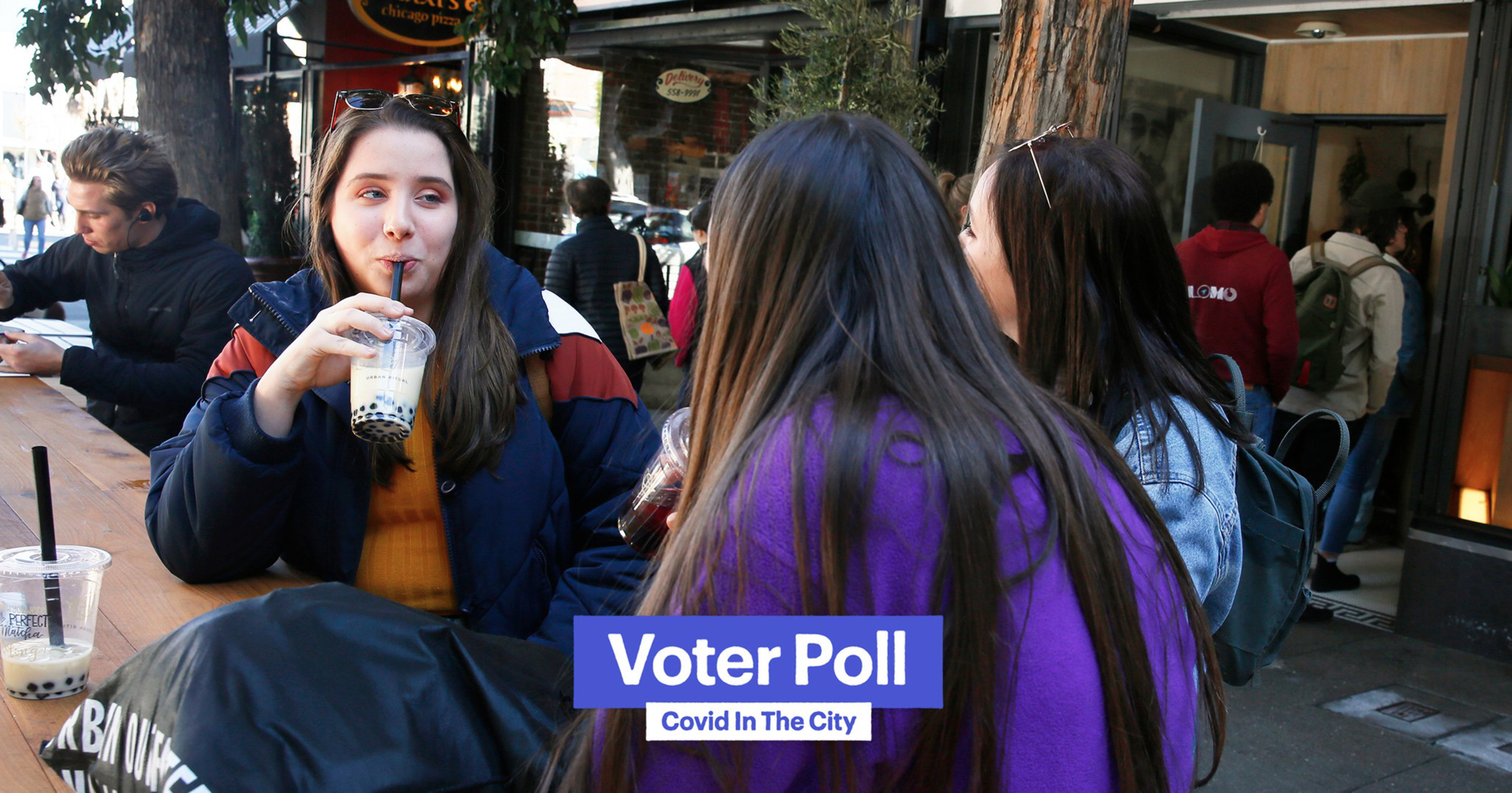 Three young women sit at an outdoor wooden table sipping bubble tea while people walk by on a city sidewalk.