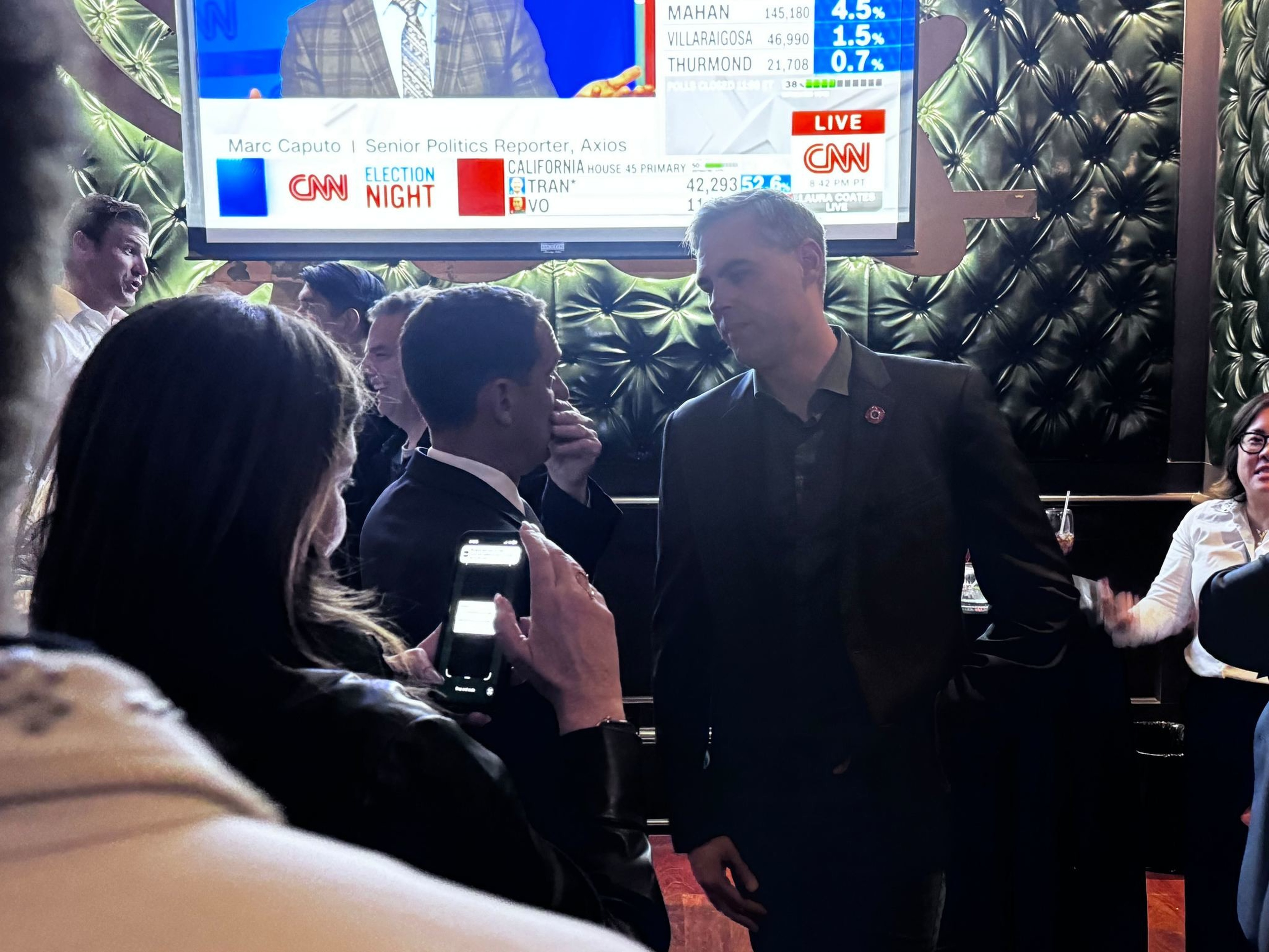 People gather indoors near a large screen showing CNN Election Night coverage, with some engaged in conversation and one person taking a photo.