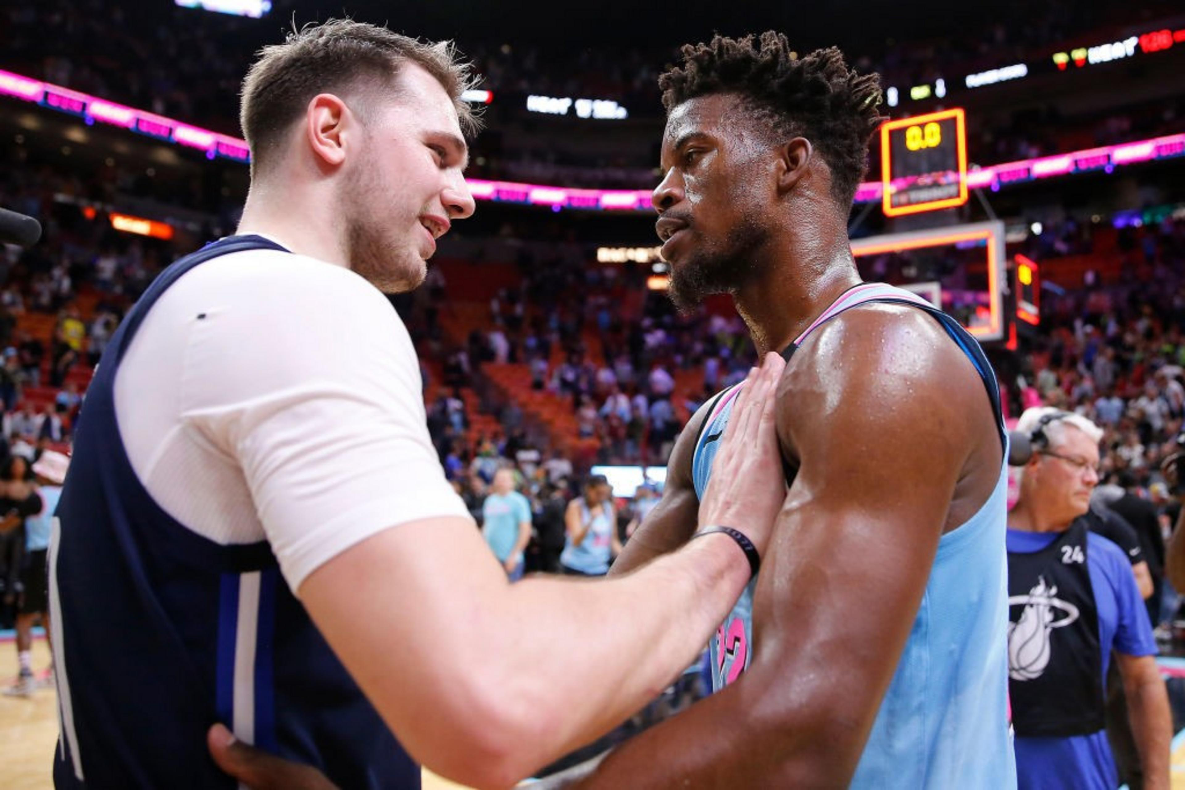 Two basketball players, one in a white and blue uniform and the other in light blue and pink, are sharing a moment on the court, surrounded by a vibrant crowd.