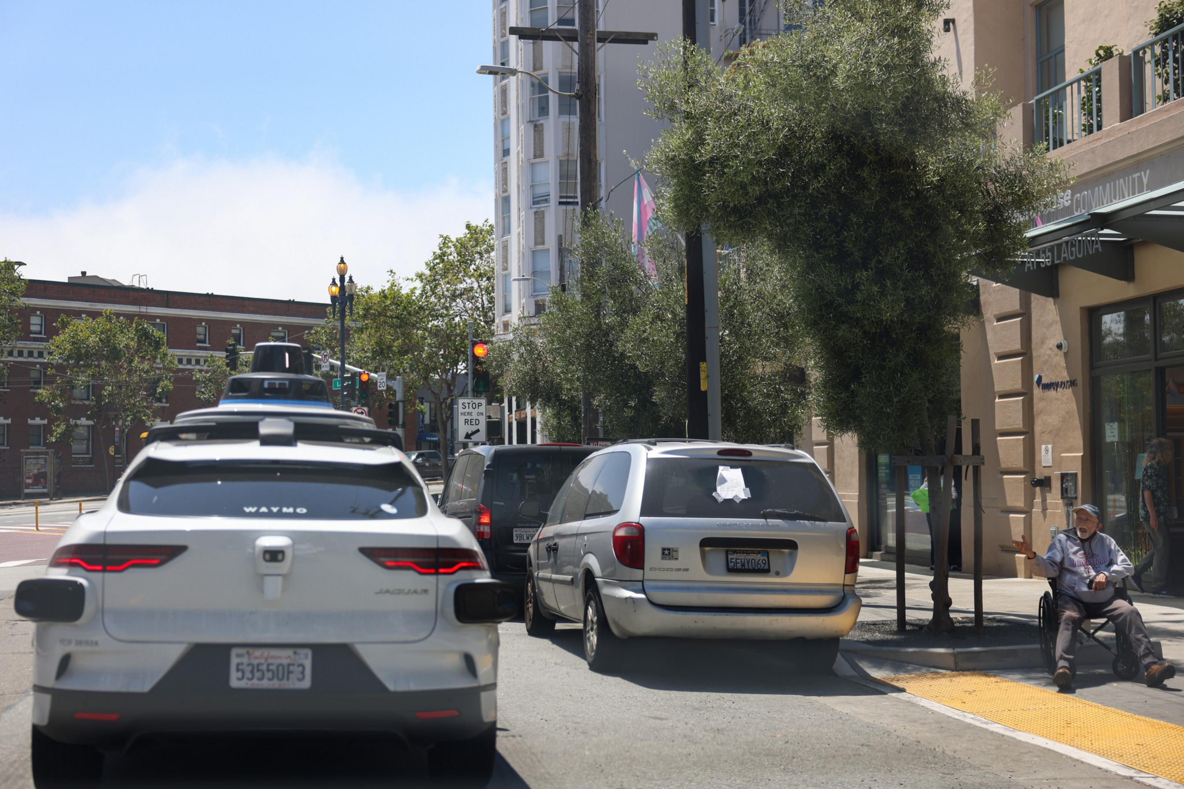 A white Waymo self-driving car and two other vehicles wait at a traffic light near a sidewalk where a person sits on the curb under trees.
