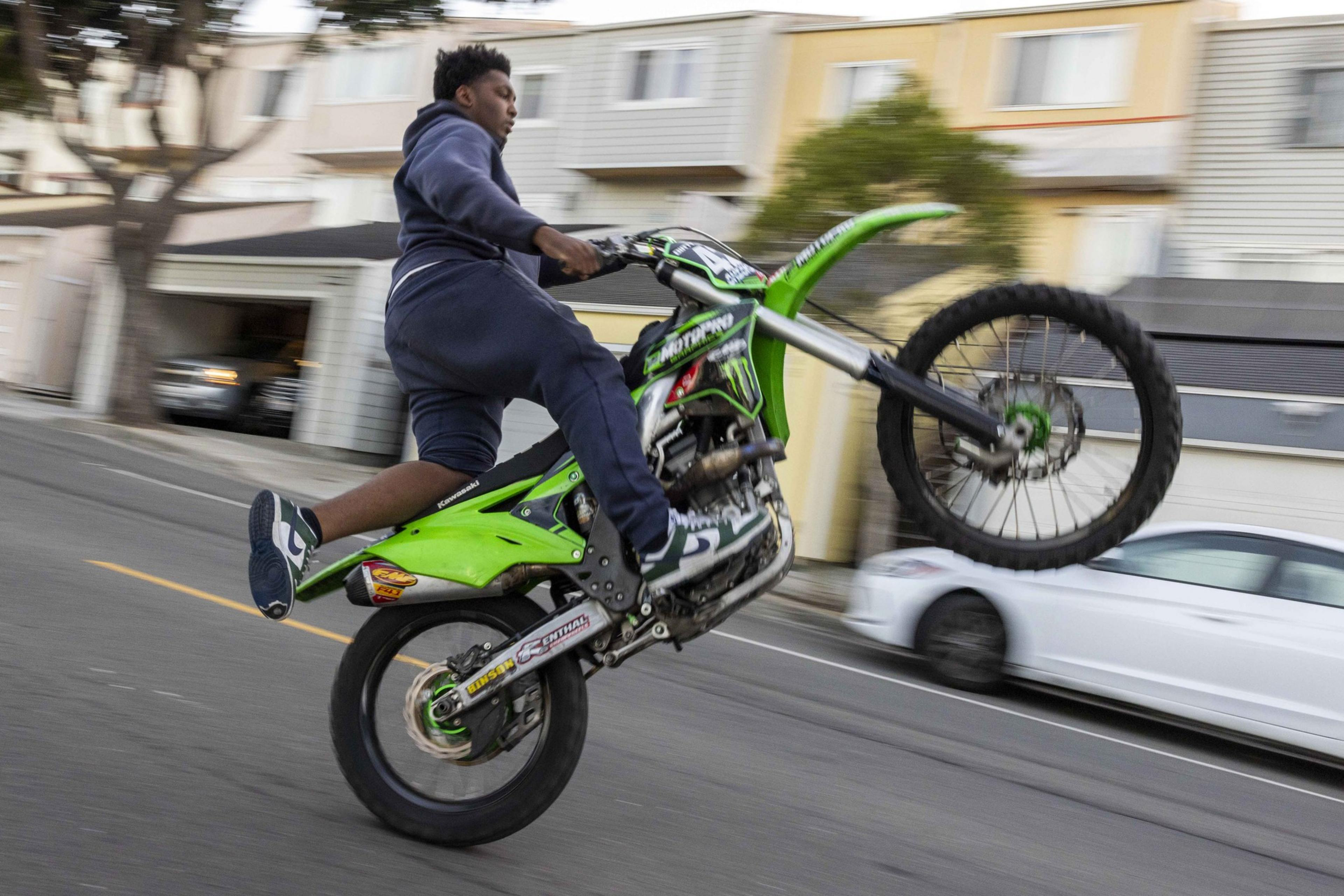A person performs a wheelie on a green motorcycle in a residential street, with houses blurred in the background, capturing motion and excitement.