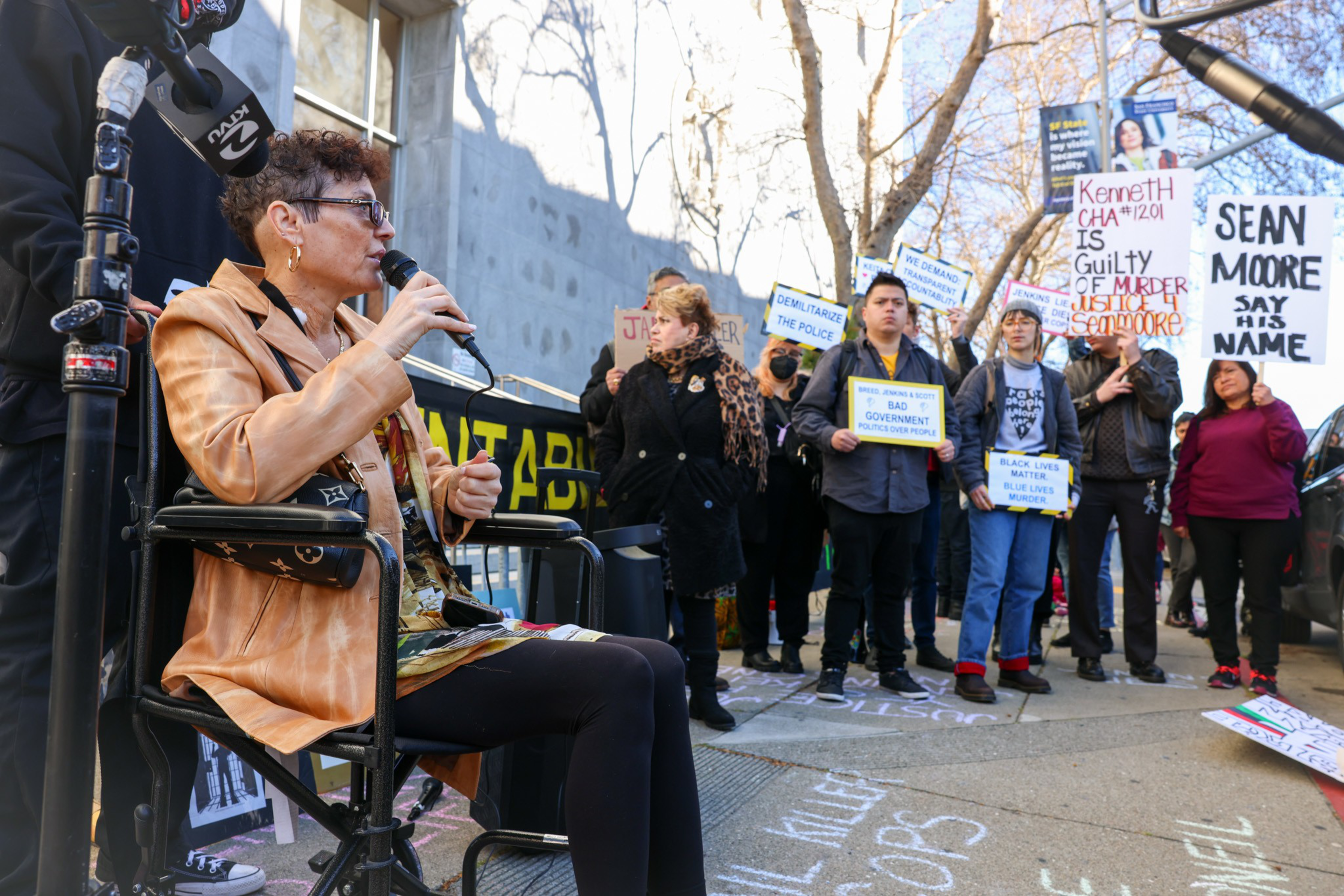 A woman in a wheelchair speaks into a microphone while a diverse group of protesters hold signs opposing police violence and injustice.