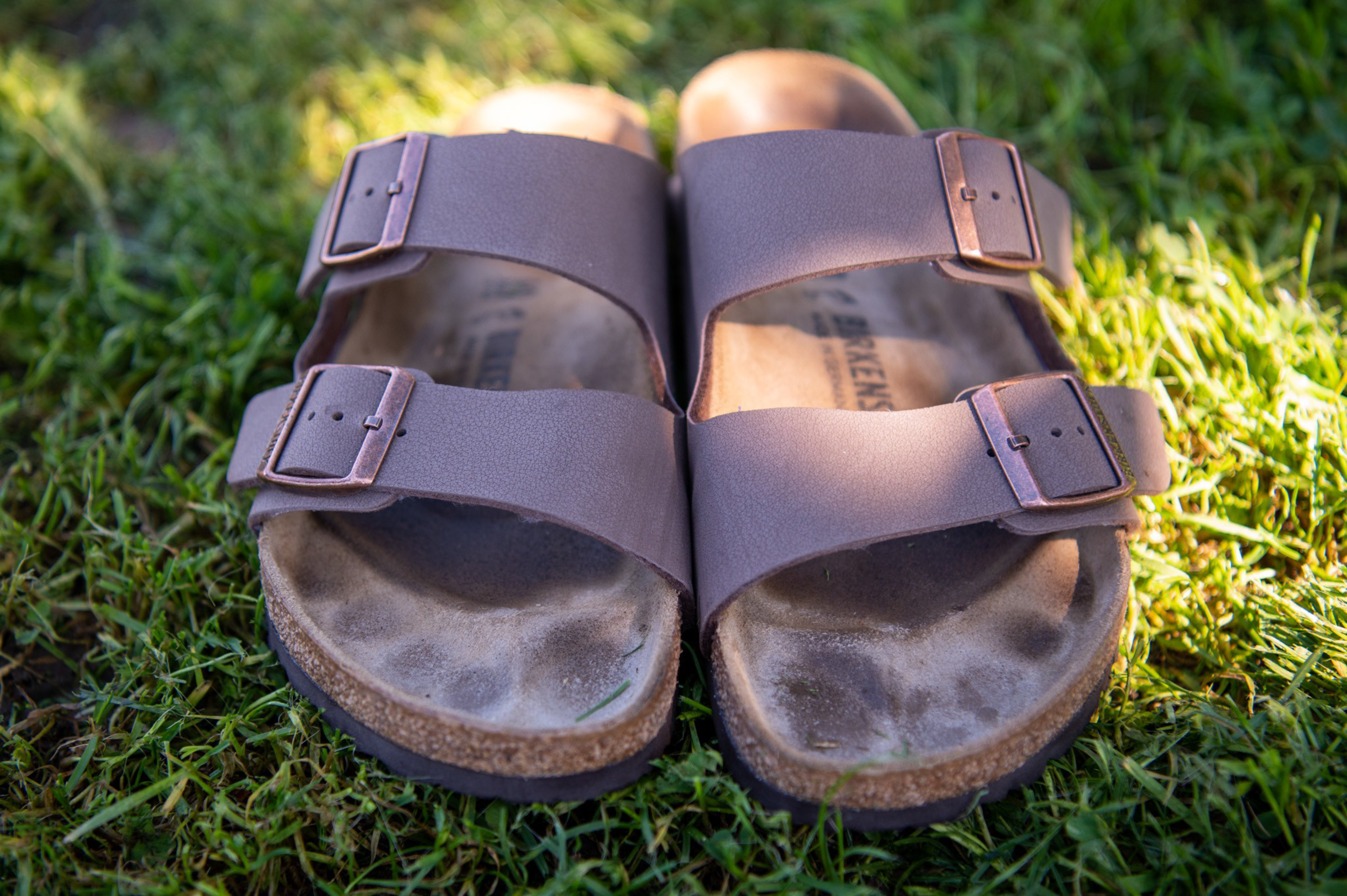 A pair of brown double-strap sandals with buckles rest on green grass, showing worn foot imprints and light dirt on the soles.