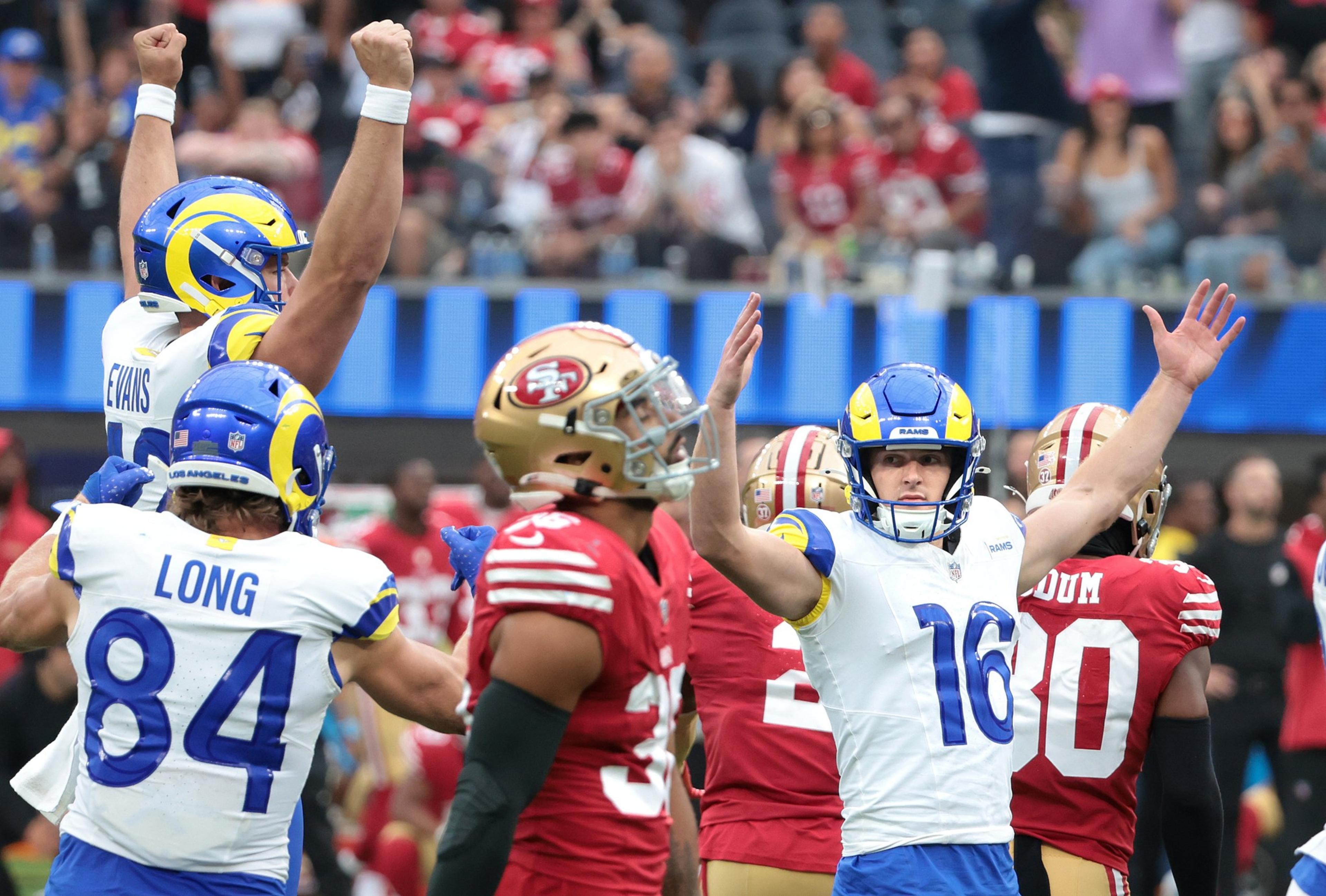 The image shows football players from the Rams celebrating with raised arms, while players from the opposing 49ers team stand nearby in red jerseys.