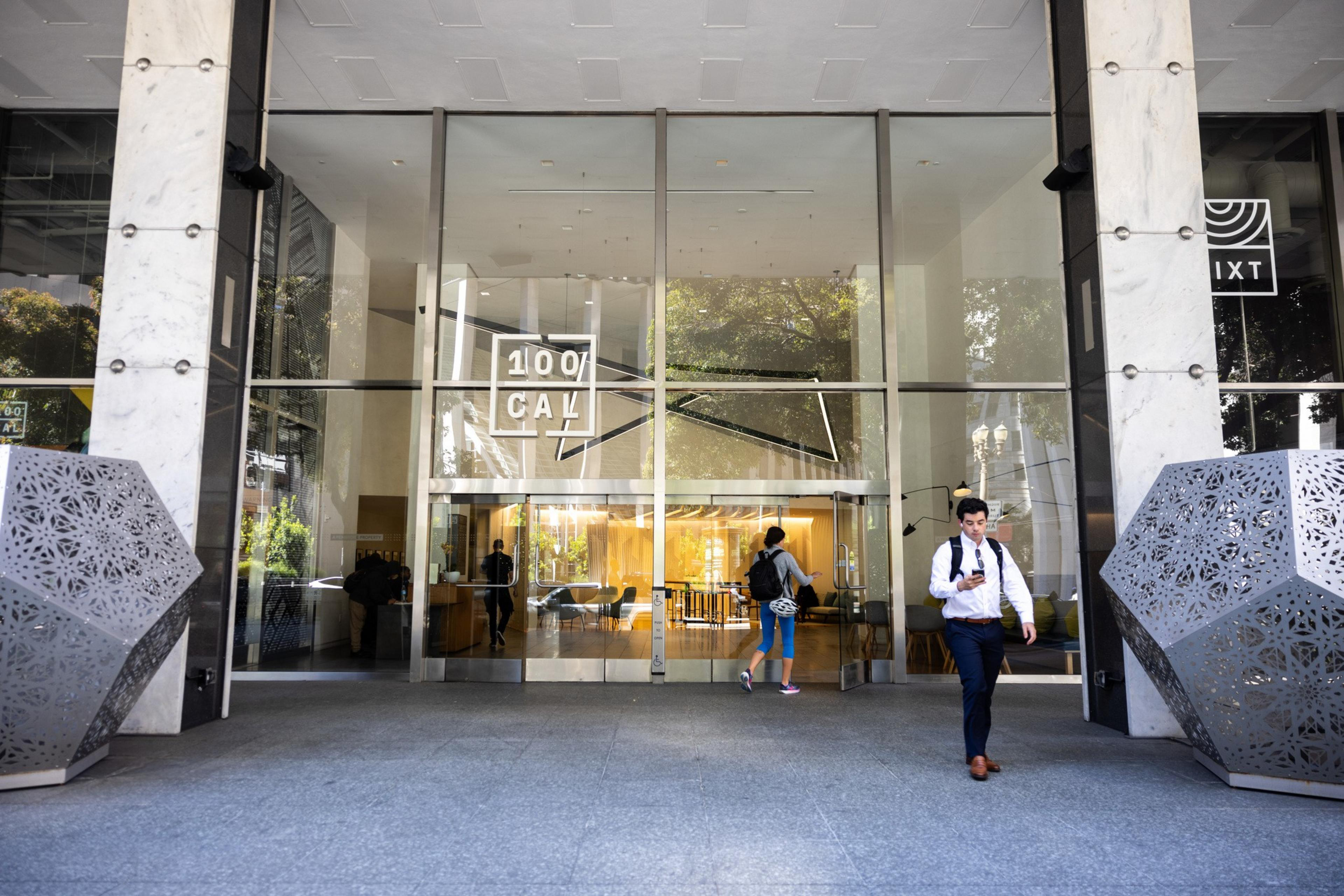 The image shows the entrance of a modern building with glass doors, labeled &quot;100 CAL,&quot; where people are entering and exiting; metal geometric sculptures enhance the scene.
