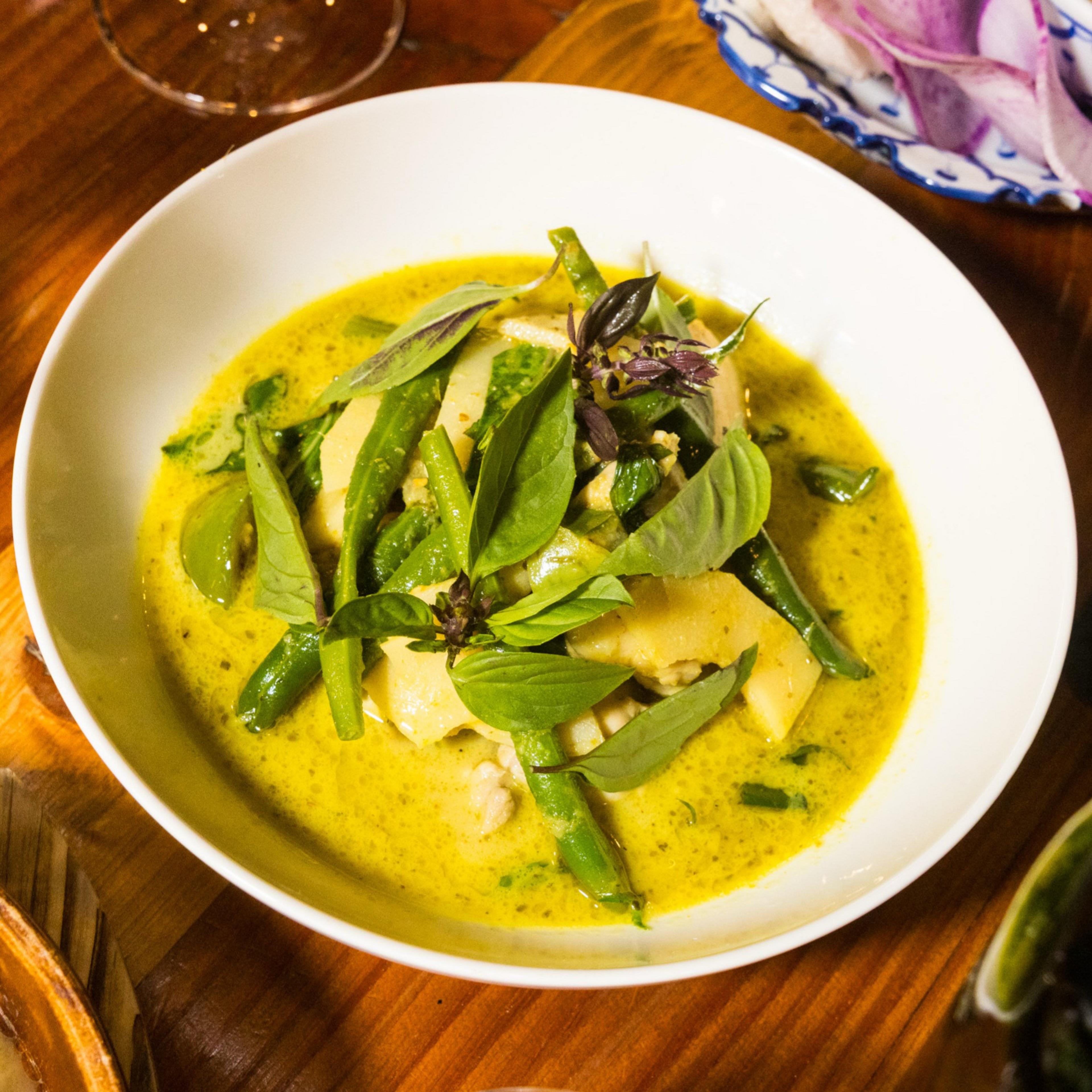 A white bowl filled with green curry, featuring visible green beans, slices of bamboo, and fresh basil leaves, sitting on a wooden table.