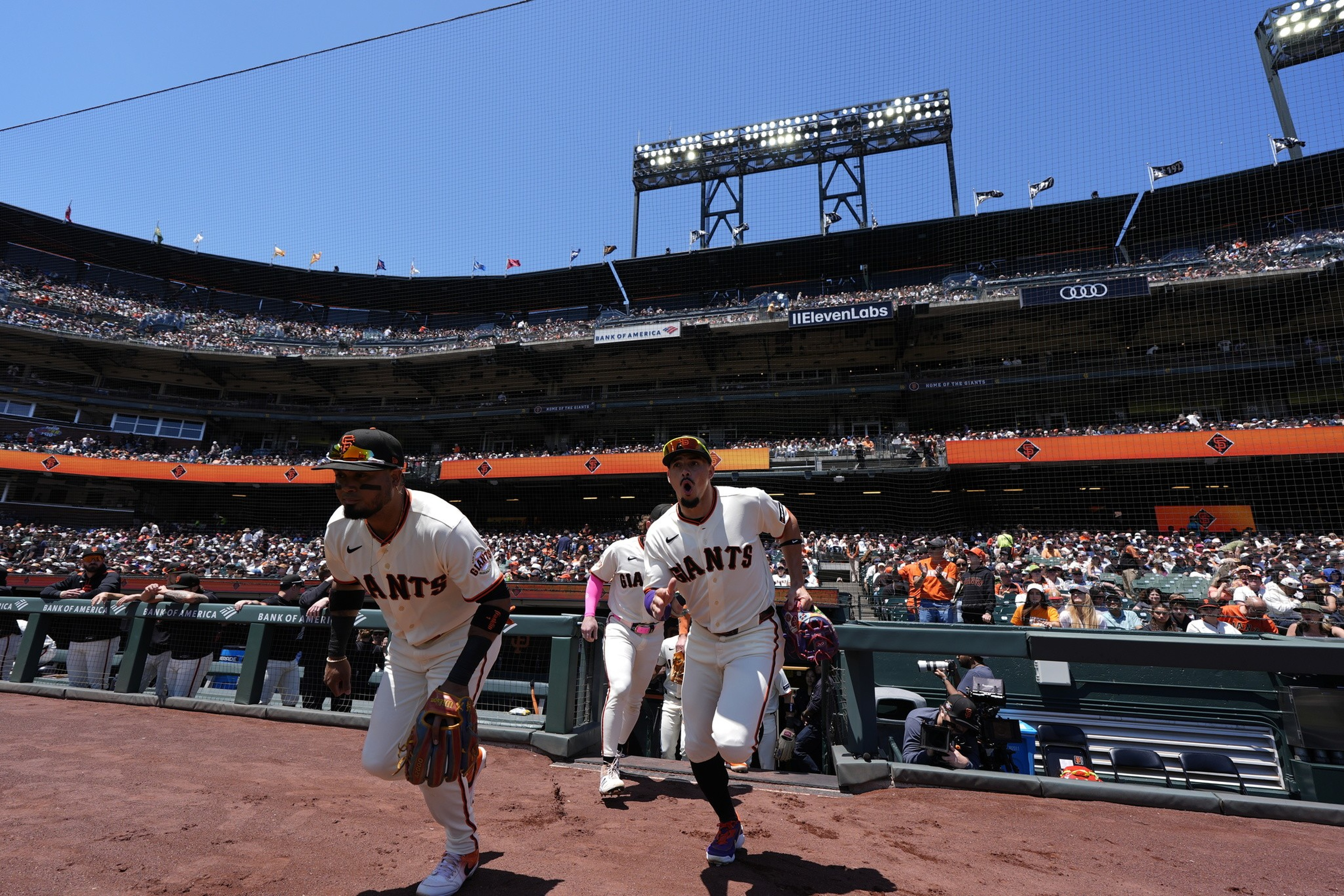 San Francisco Giants baseball players are running onto the field from the dugout with a large crowd watching in a sunny stadium.