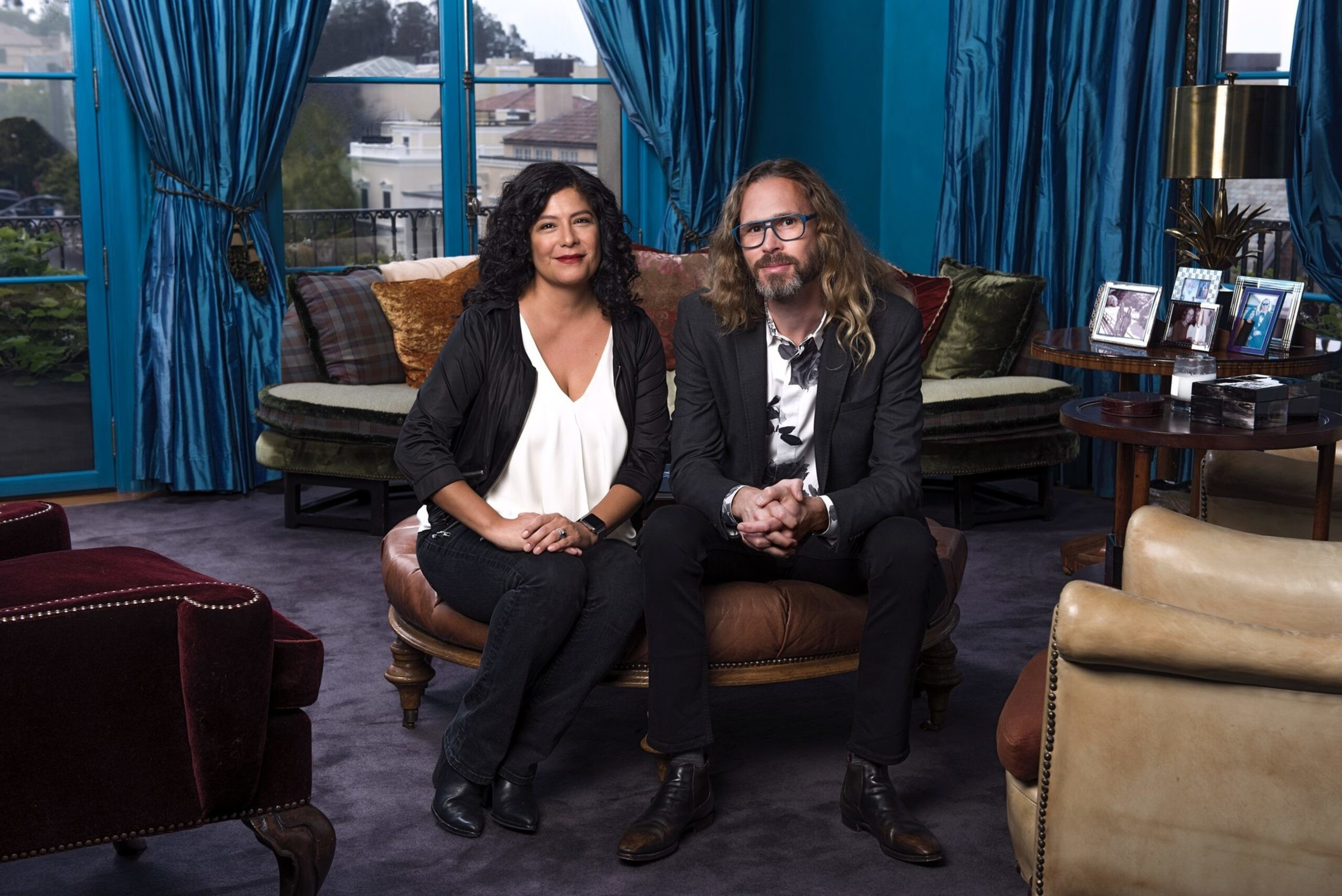 A woman with curly dark hair and a man with long blond hair sit side by side on a leather ottoman in a room with teal curtains and framed photos.