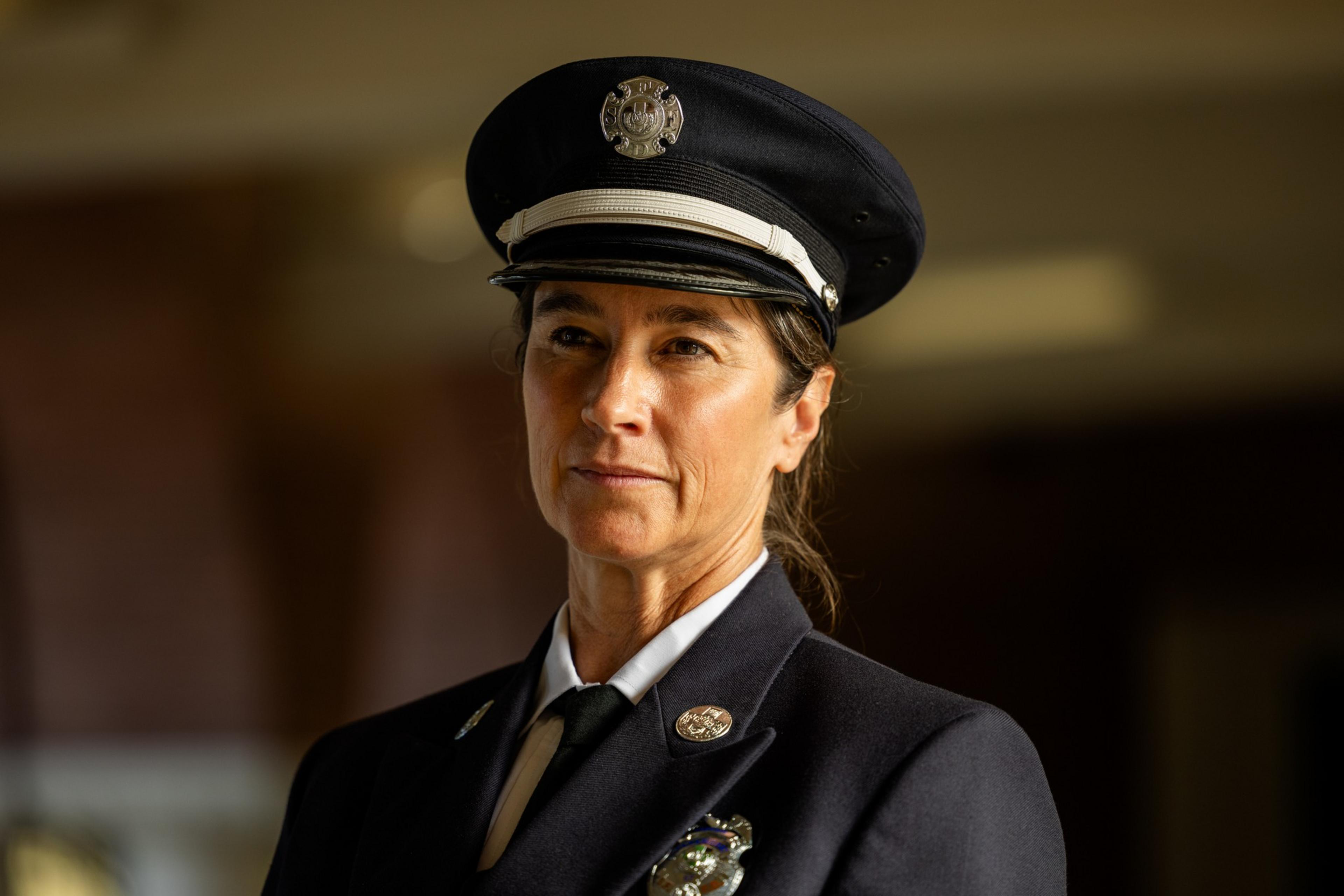 A middle-aged woman wearing a dark firefighter dress uniform and cap looks slightly to the side with a calm, confident expression.