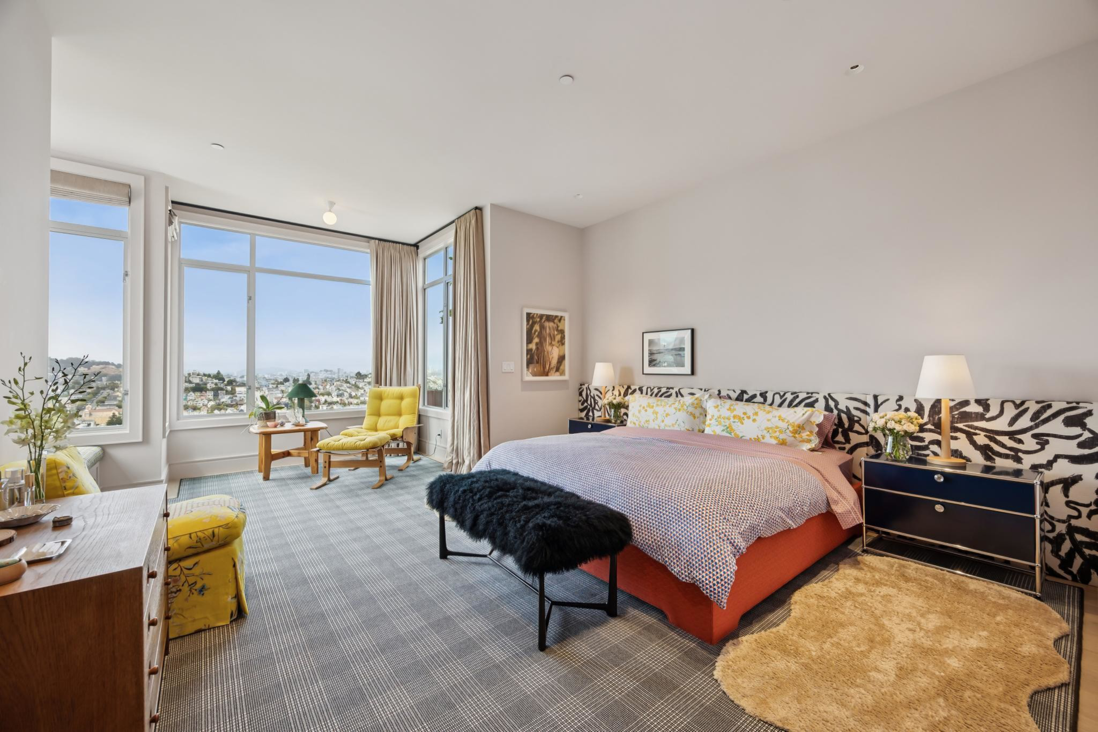 A spacious bedroom features a large window with city views, a bed with patterned headboard, yellow chairs, black bench, and light brown rug.