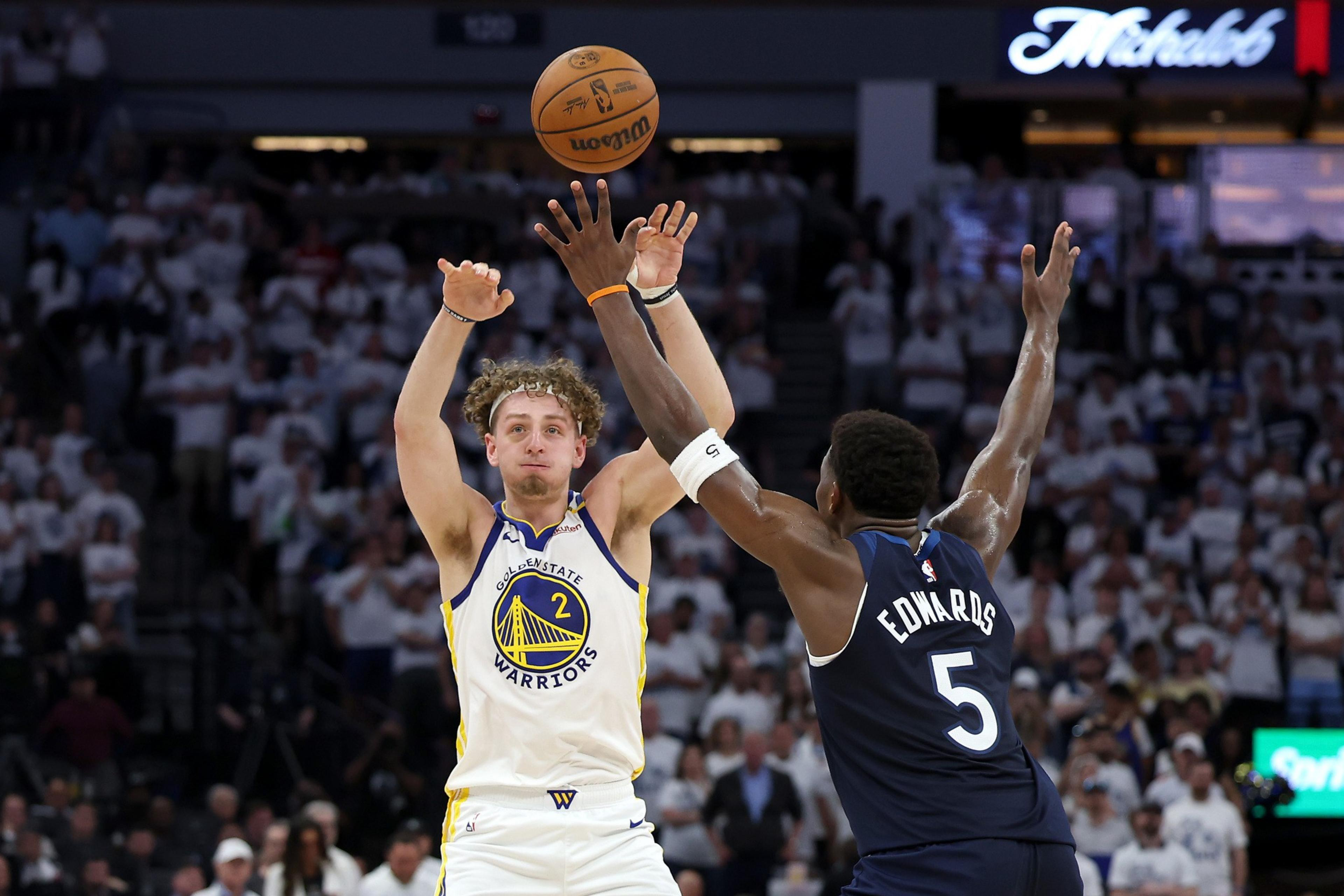 A basketball player in a white jersey passes the ball over a player's outstretched arms in a dark jersey during a game, with a crowd in the background.