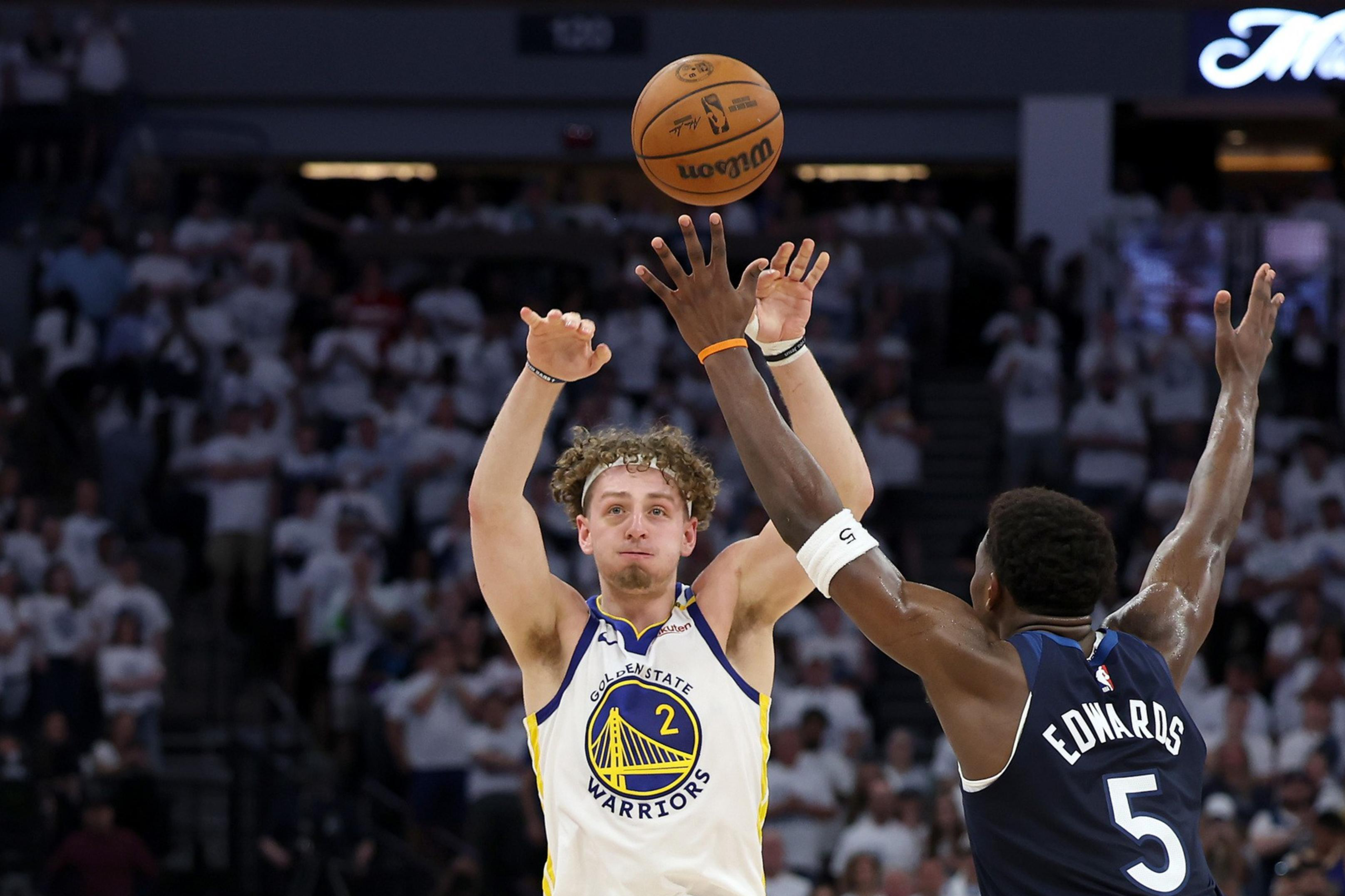 A basketball player in a white jersey passes the ball over a player's outstretched arms in a dark jersey during a game, with a crowd in the background.
