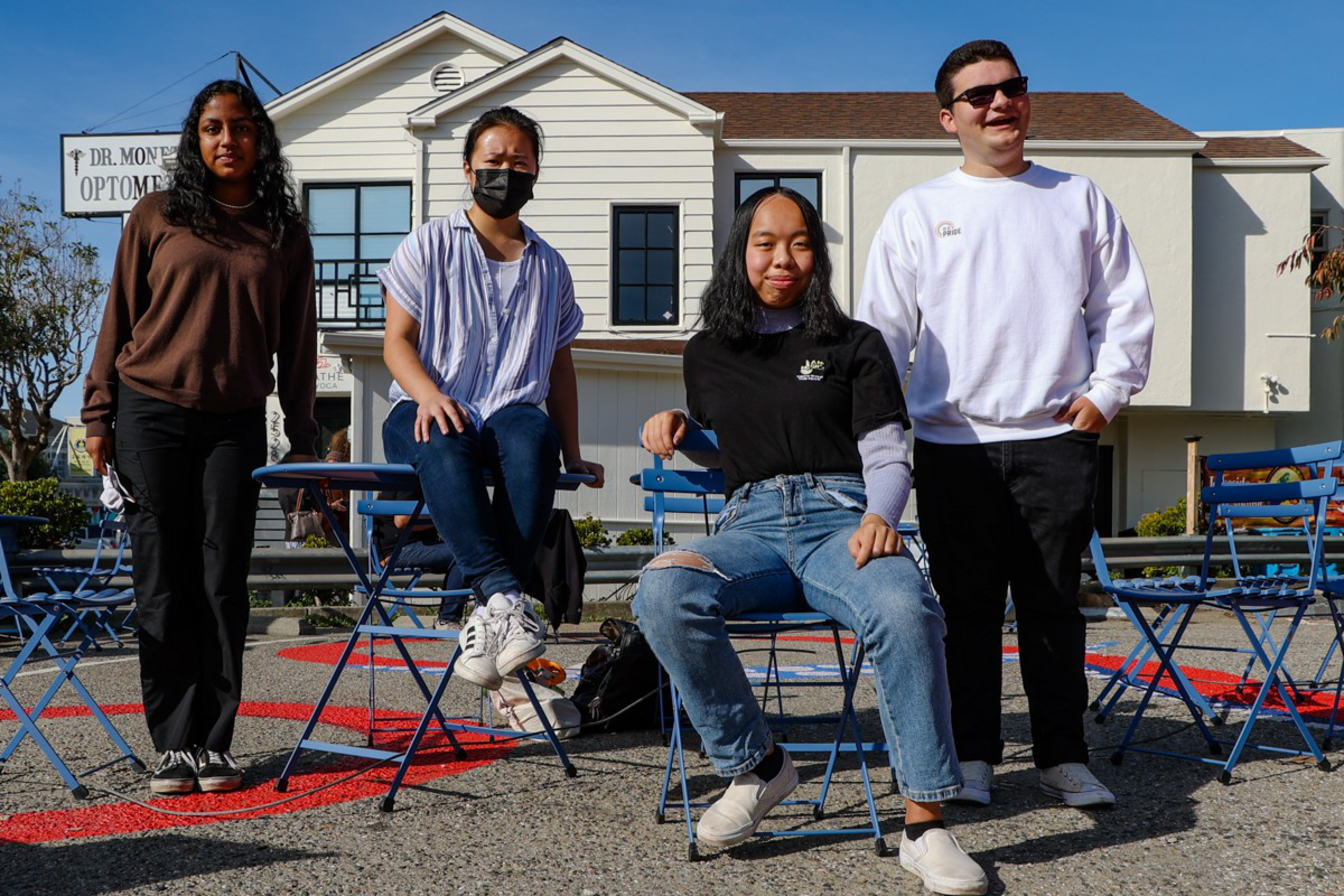 Four people pose with blue metal chairs and tables on a sunny outdoor pavement in front of a white building with an "Optometry" sign.