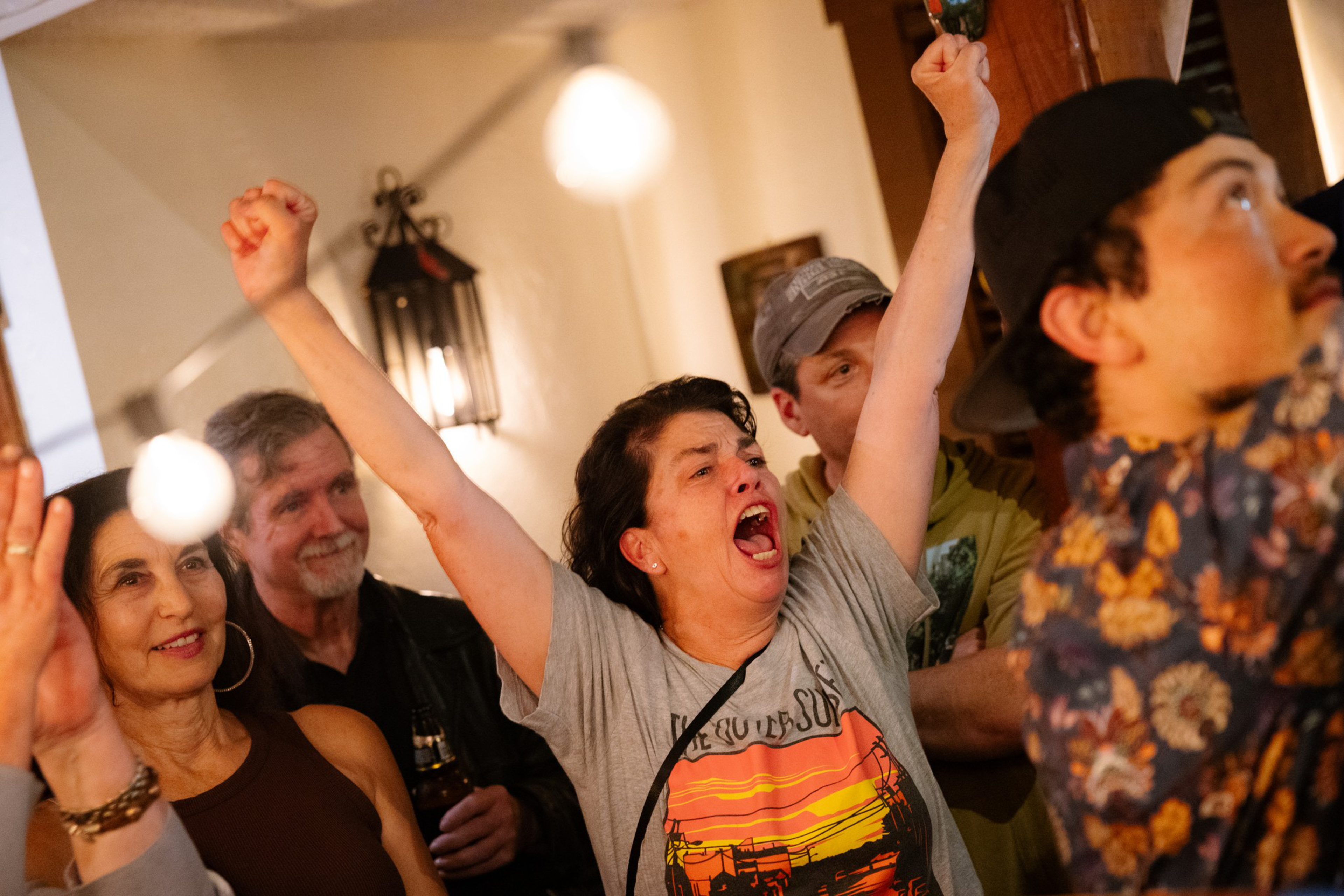 A woman in a gray shirt raises her arms and shouts joyfully while others around her smile and watch attentively in a warmly lit room.