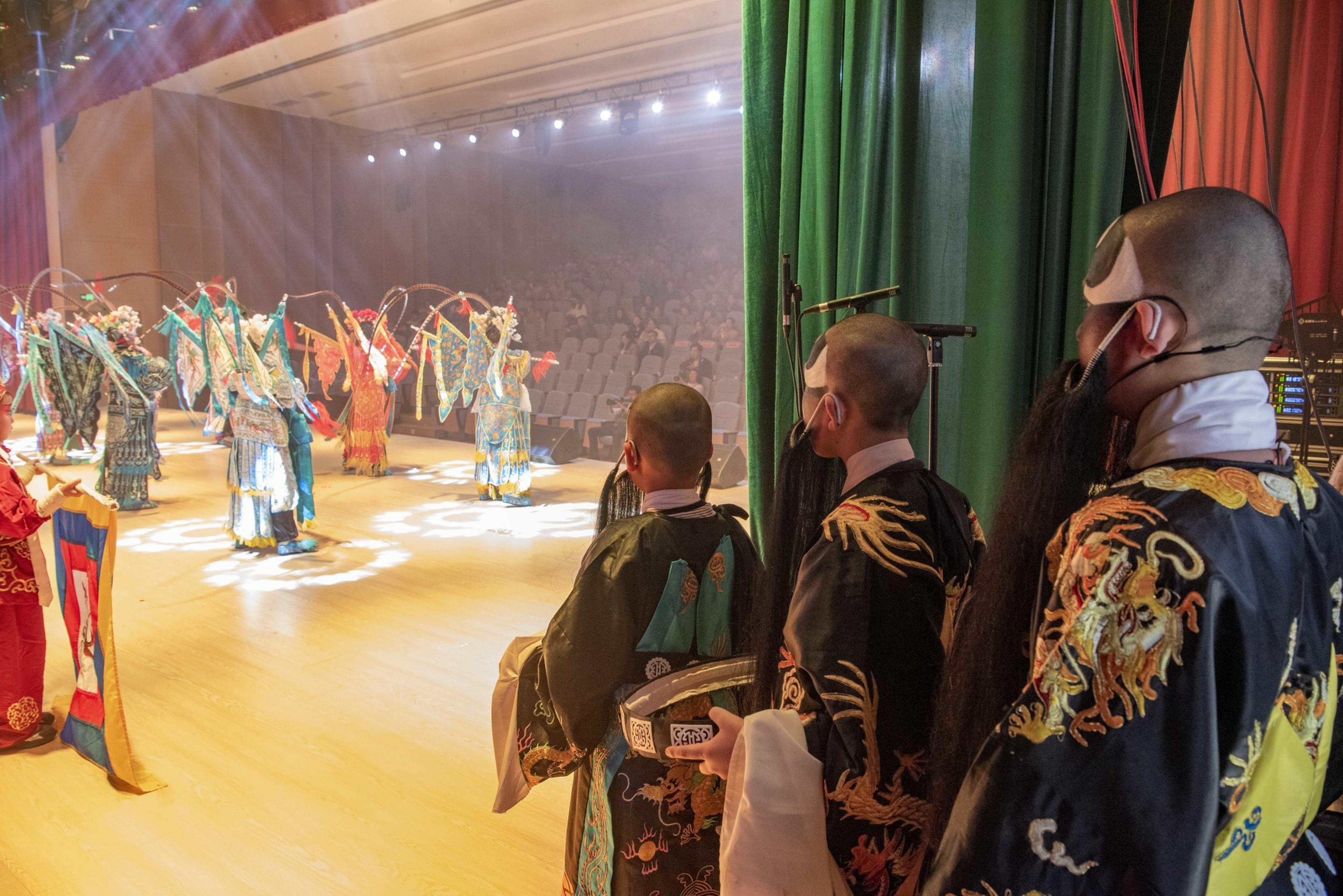 Performers backstage, wearing ornate costumes and masks, observe a vibrant, costumed dance on a brightly lit stage with an audience seated in the background.
