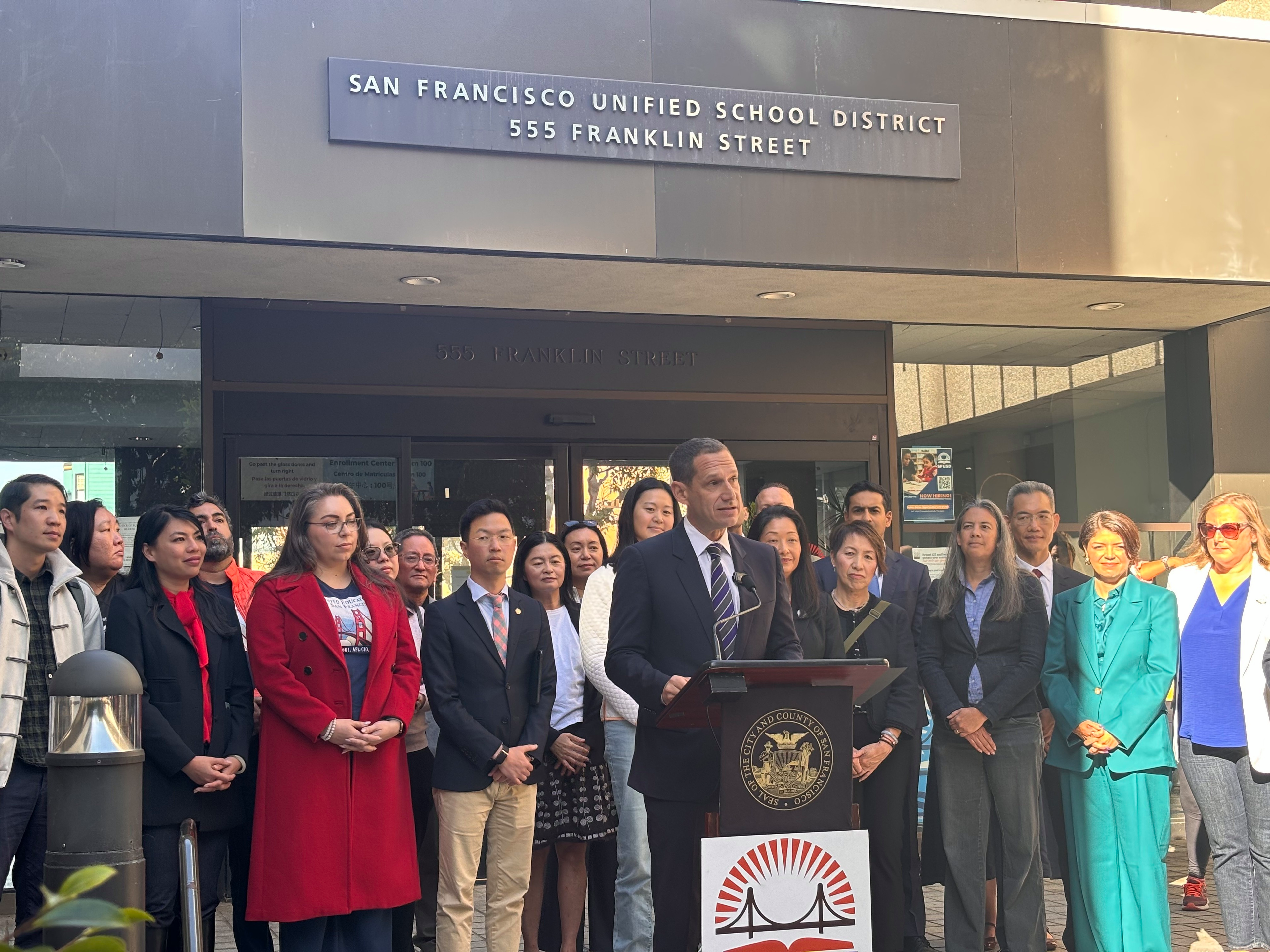 A man in a suit speaks at a podium outside San Francisco Unified School District, surrounded by a diverse group of attentive people.