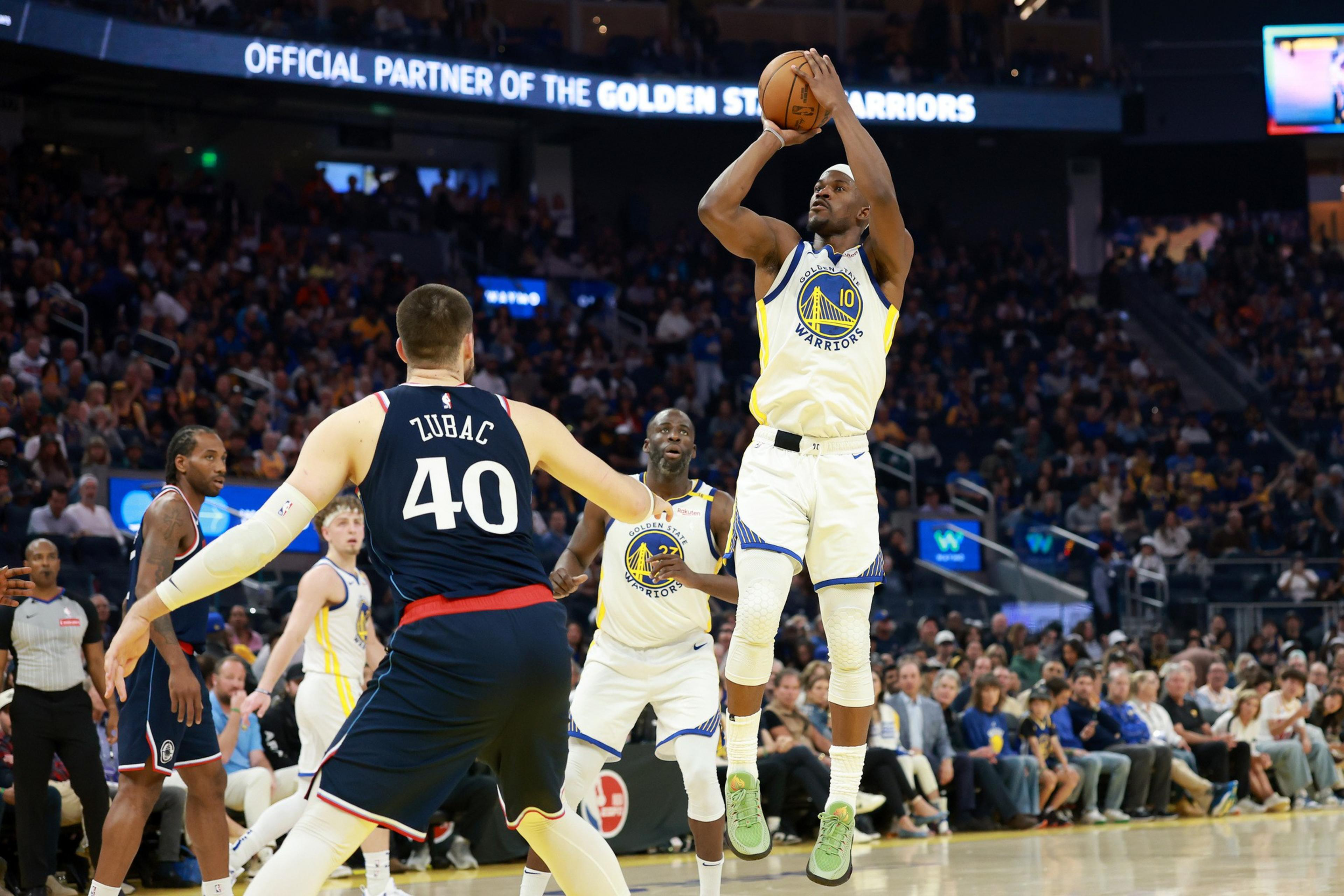 A basketball player wearing a Golden State Warriors jersey jumps to shoot the ball, while an opposing player defends. The crowd watches intently.