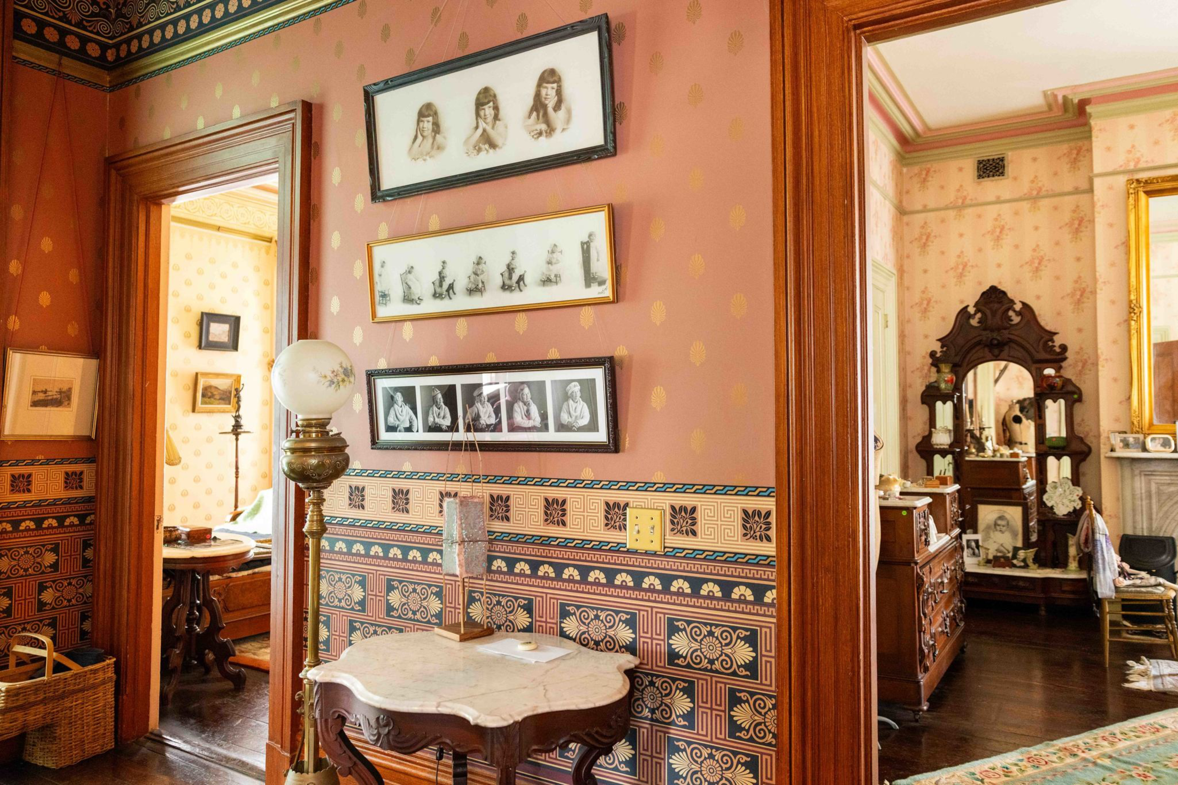 A vintage room features patterned wallpaper, framed black-and-white portraits, a marble-top table with a decorative lamp, and wooden door frames.