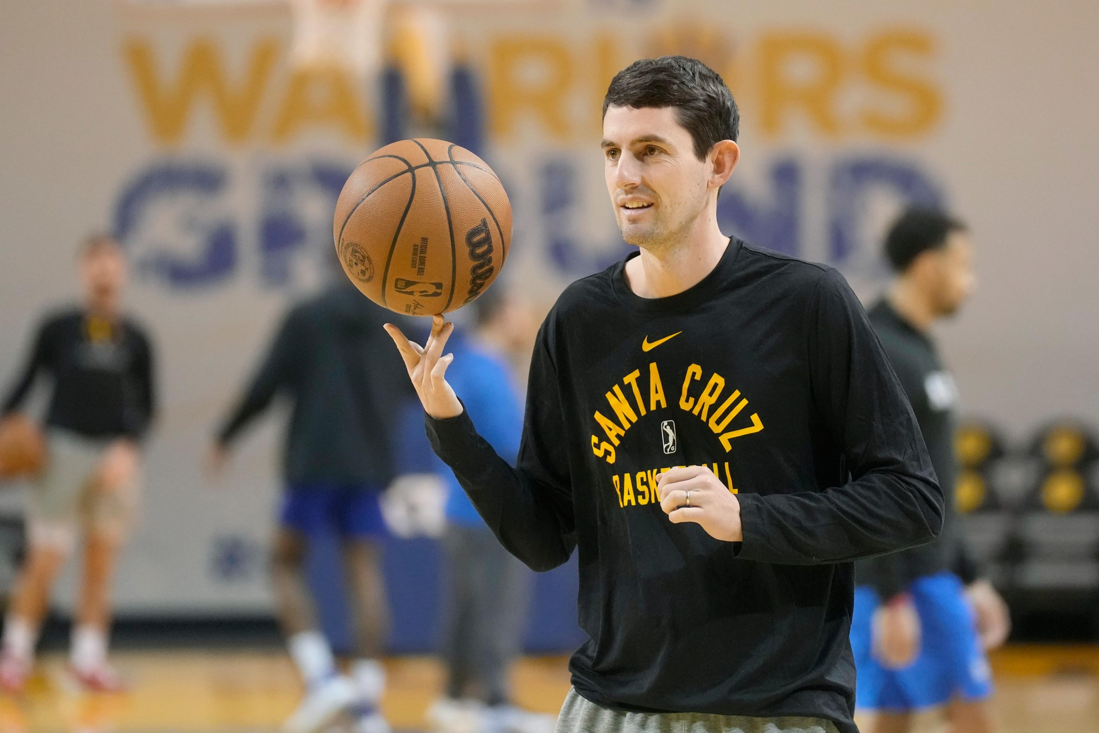 A man in a black "Santa Cruz Basketball" shirt spins a basketball on his finger in a gym with blurred people and a "Warriors" sign in the background.