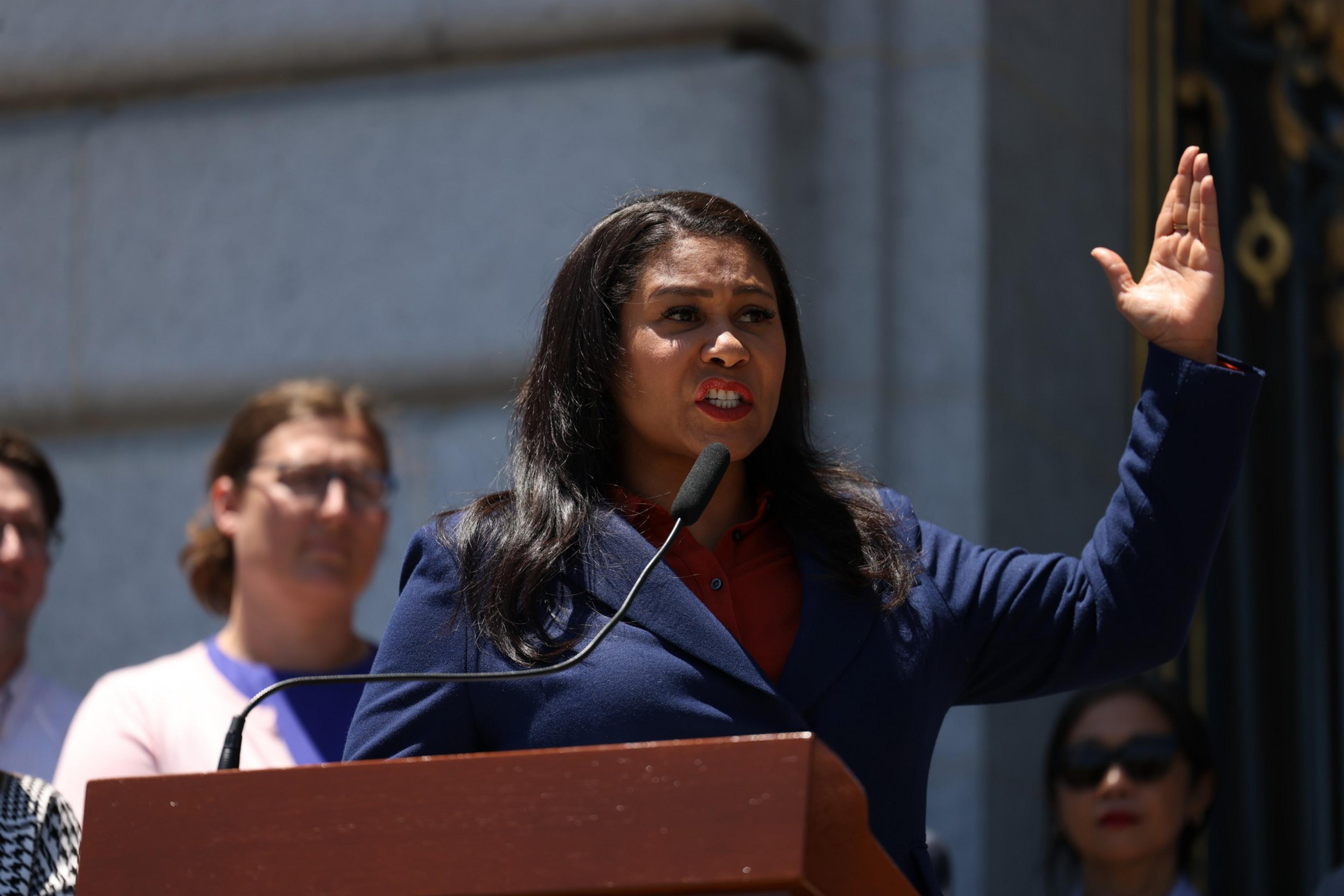 A woman in a navy blazer speaks passionately at a podium with a microphone, raising her right hand while people stand blurred behind her.