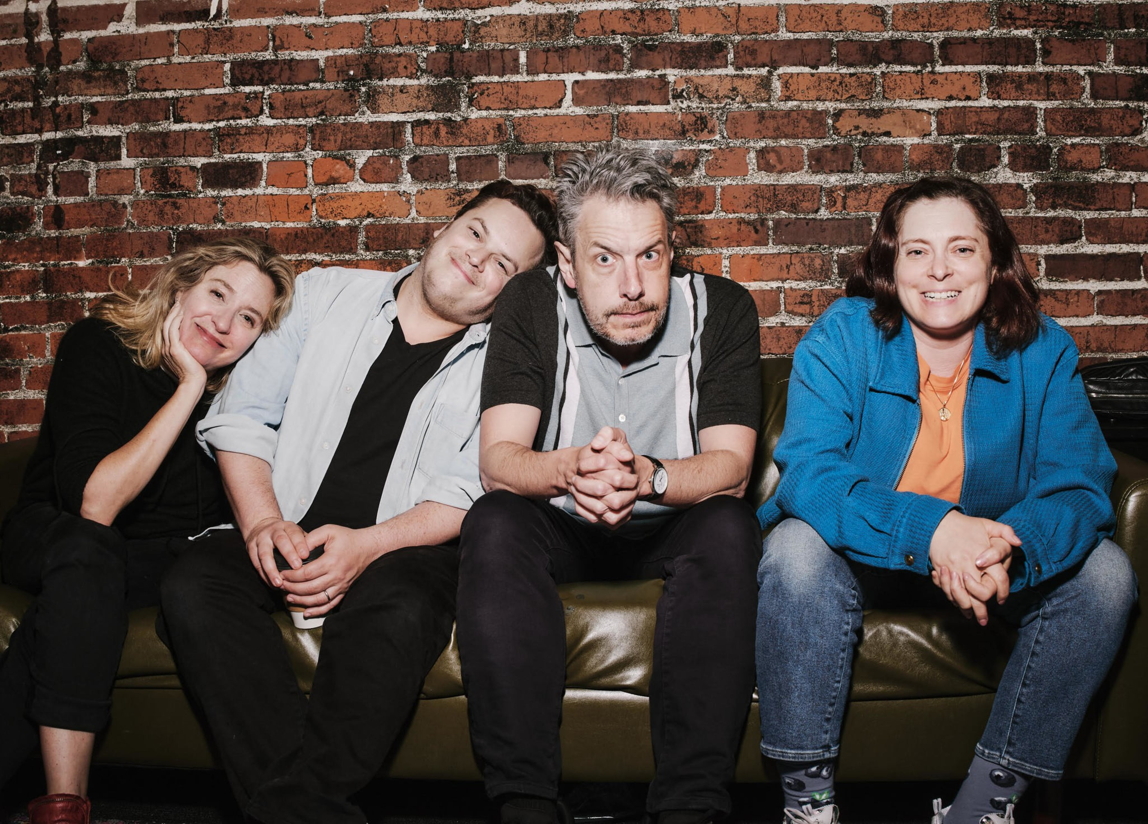 Four people sit comfortably on a green couch in front of a brick wall, smiling and leaning on each other. One wears a blue jacket, another a striped shirt.