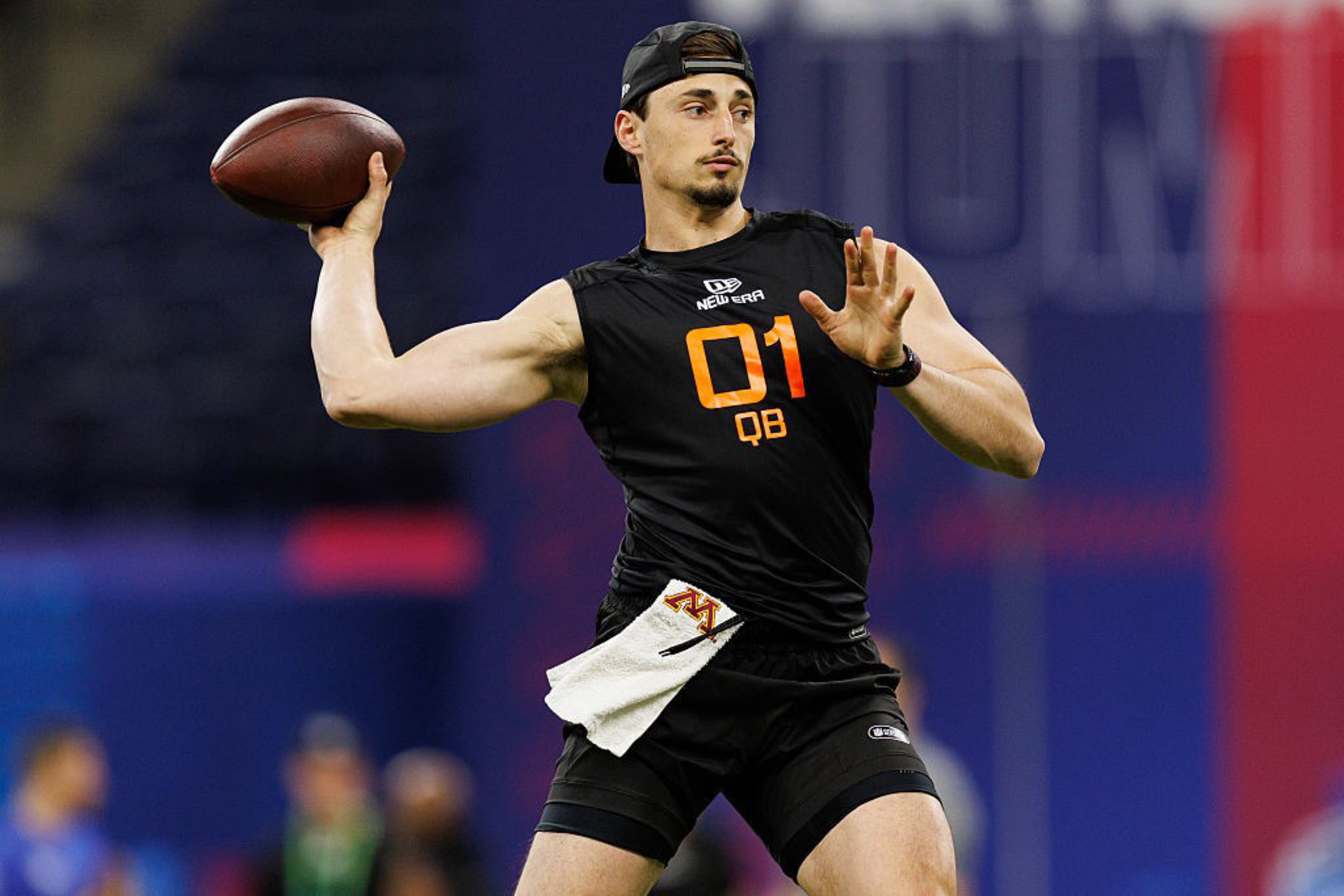 A person in a black athletic outfit with &quot;01 QB&quot; written on it is poised to throw a football. They wear a backward cap and have a towel tucked into their waistband.