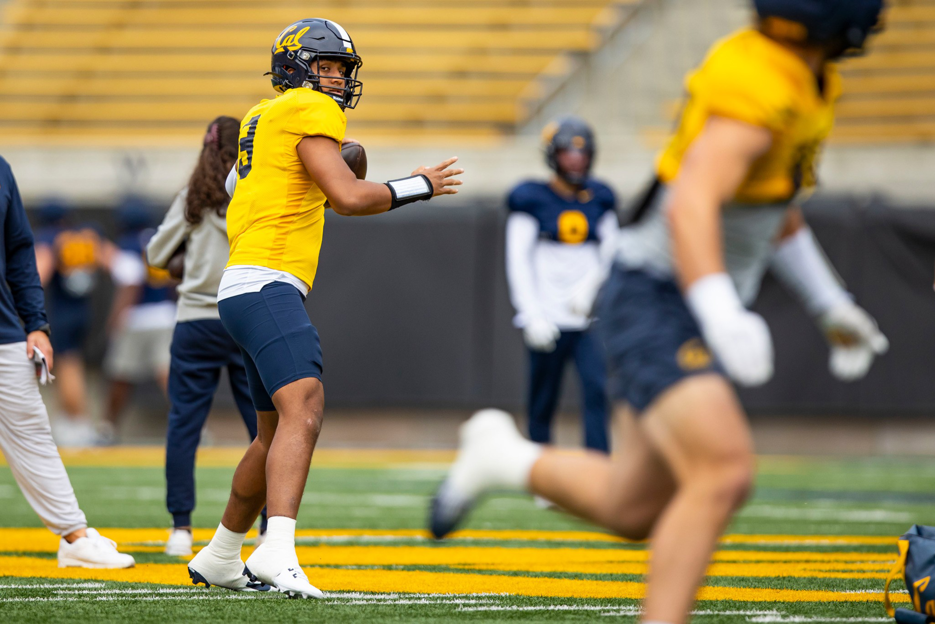 A football player in a yellow jersey and helmet prepares to throw a ball while another player runs on the field, with teammates and empty stands in the background.