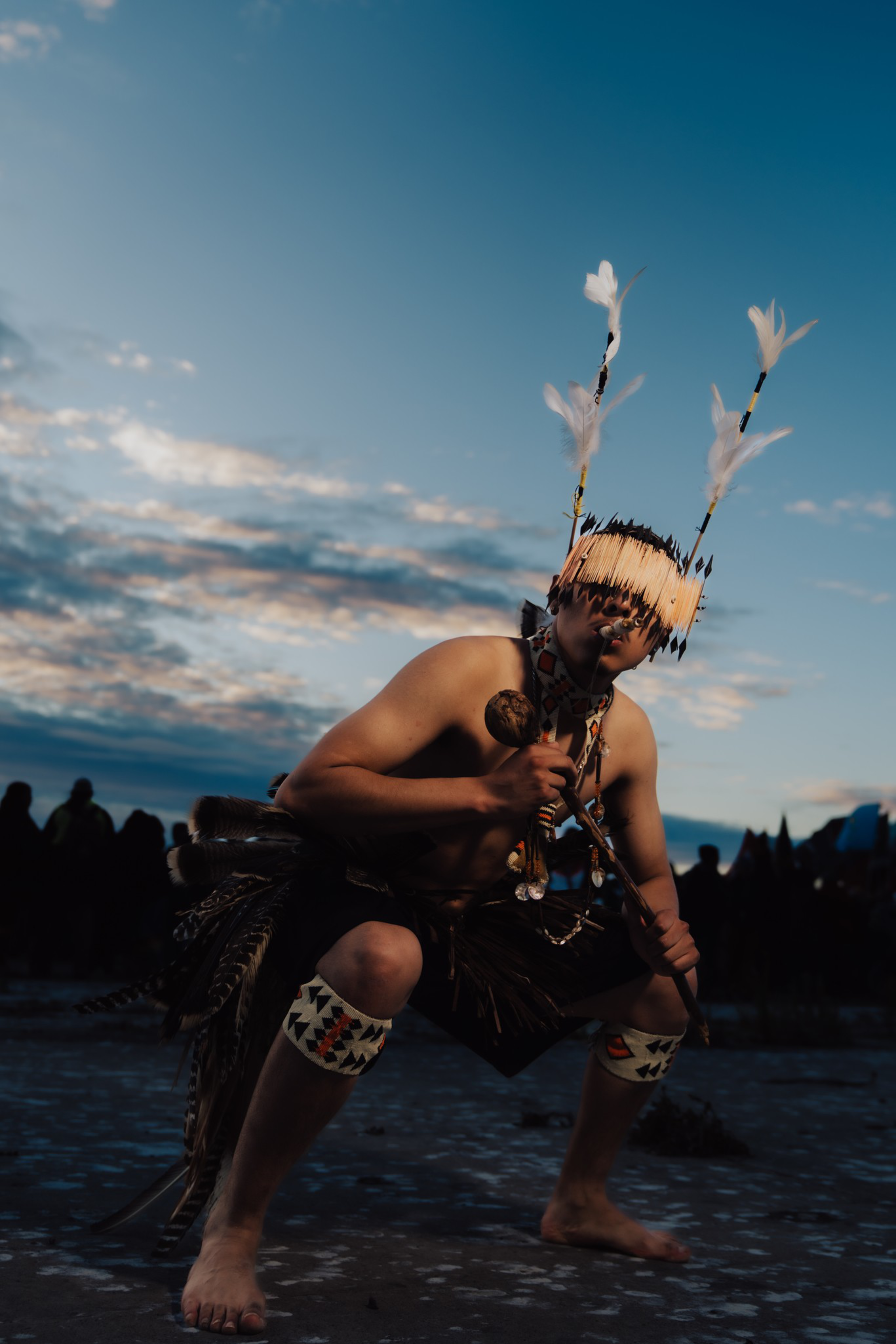 A person wearing traditional regalia with feathered headgear crouches on rocky ground under a twilight sky.