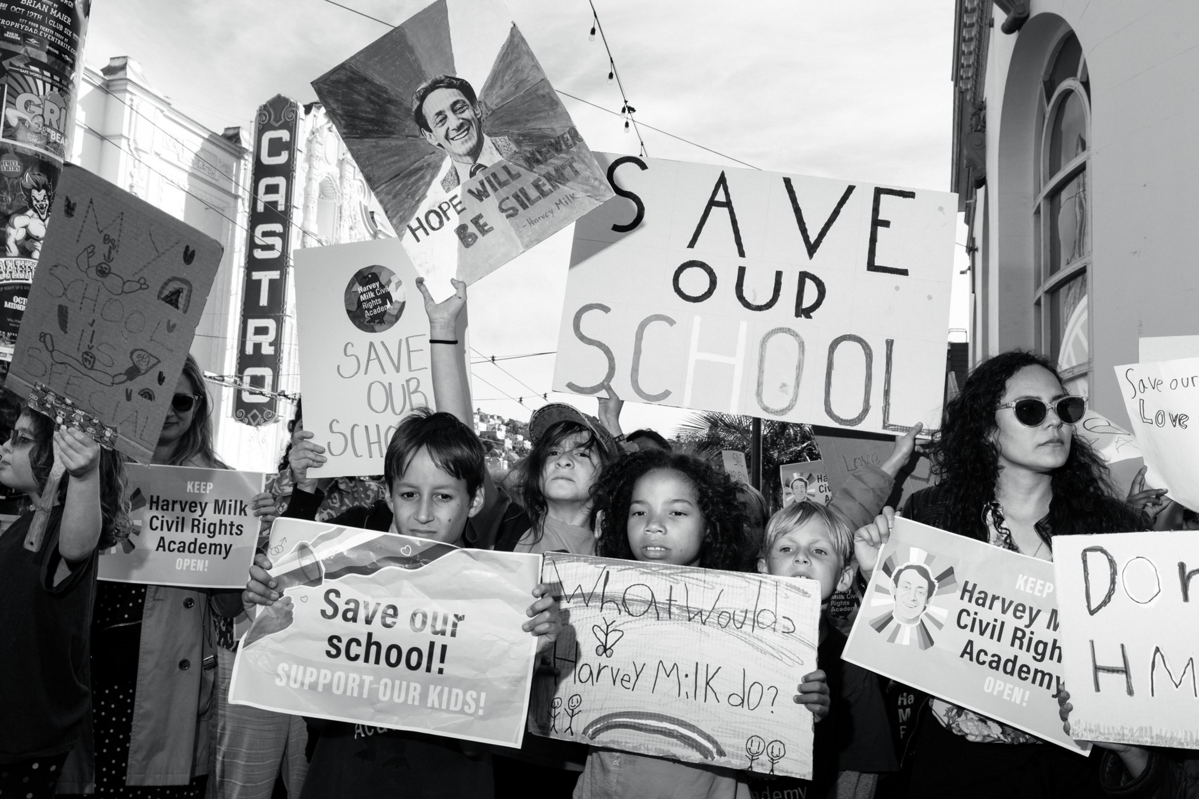 A group of people, including children, hold signs in protest reading "Save Our School" and "Keep Harvey Milk Civil Rights Academy Open!" in a busy street scene.