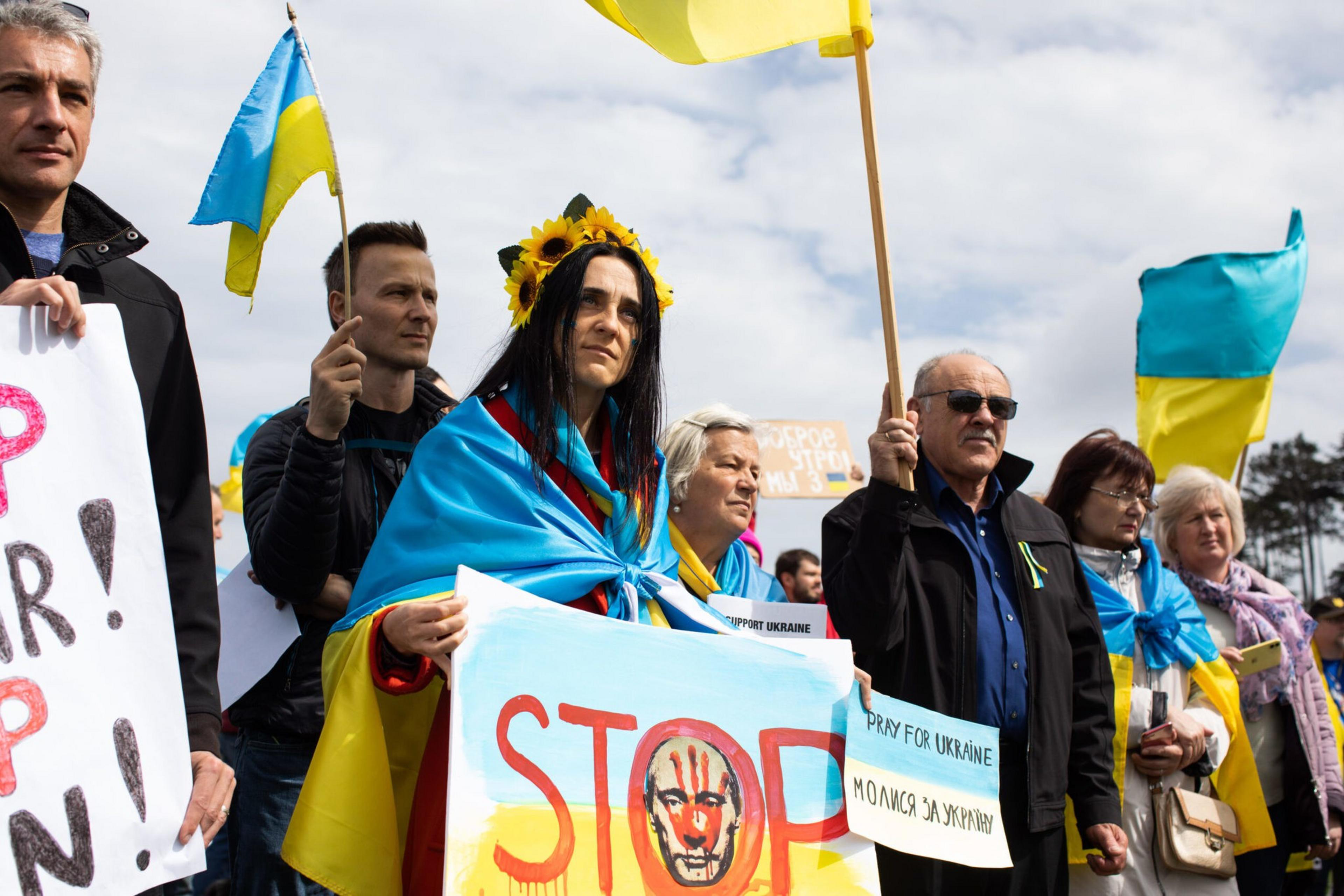 People stand together holding Ukrainian flags, wearing blue and yellow with one woman wearing a sunflower crown, holding a sign that says "STOP" and "PRAY FOR UKRAINE."