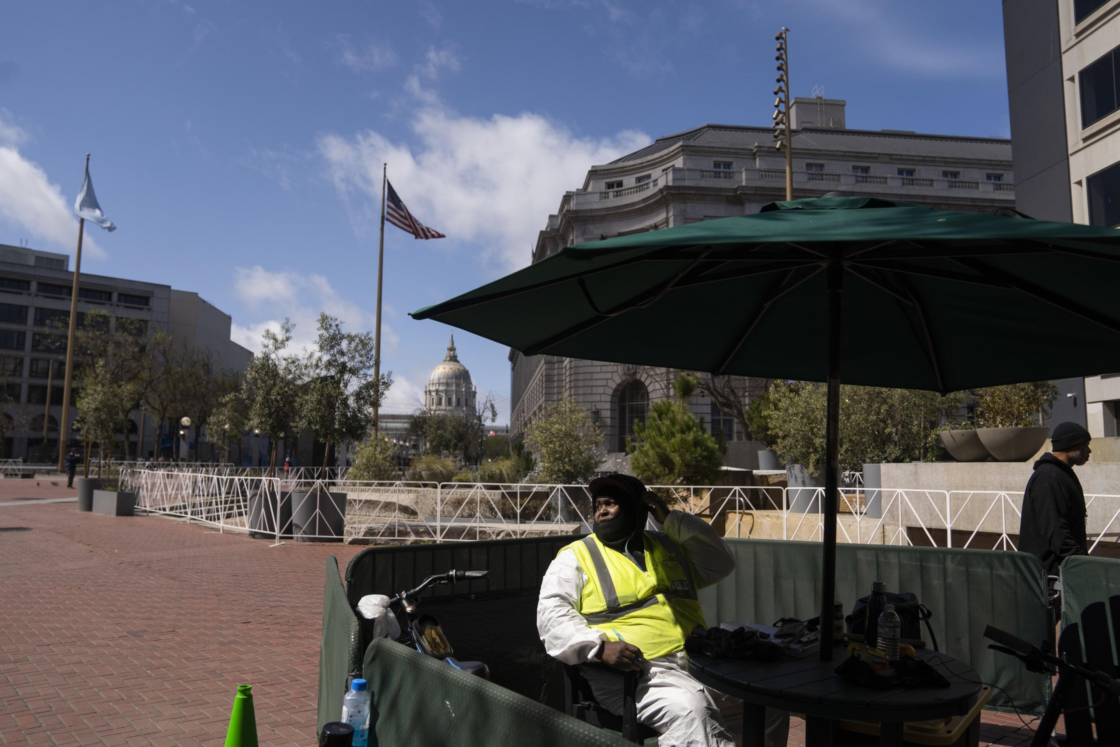 A person in a yellow safety vest sits under a green umbrella, surrounded by a partition, with a city building and flag in the background.