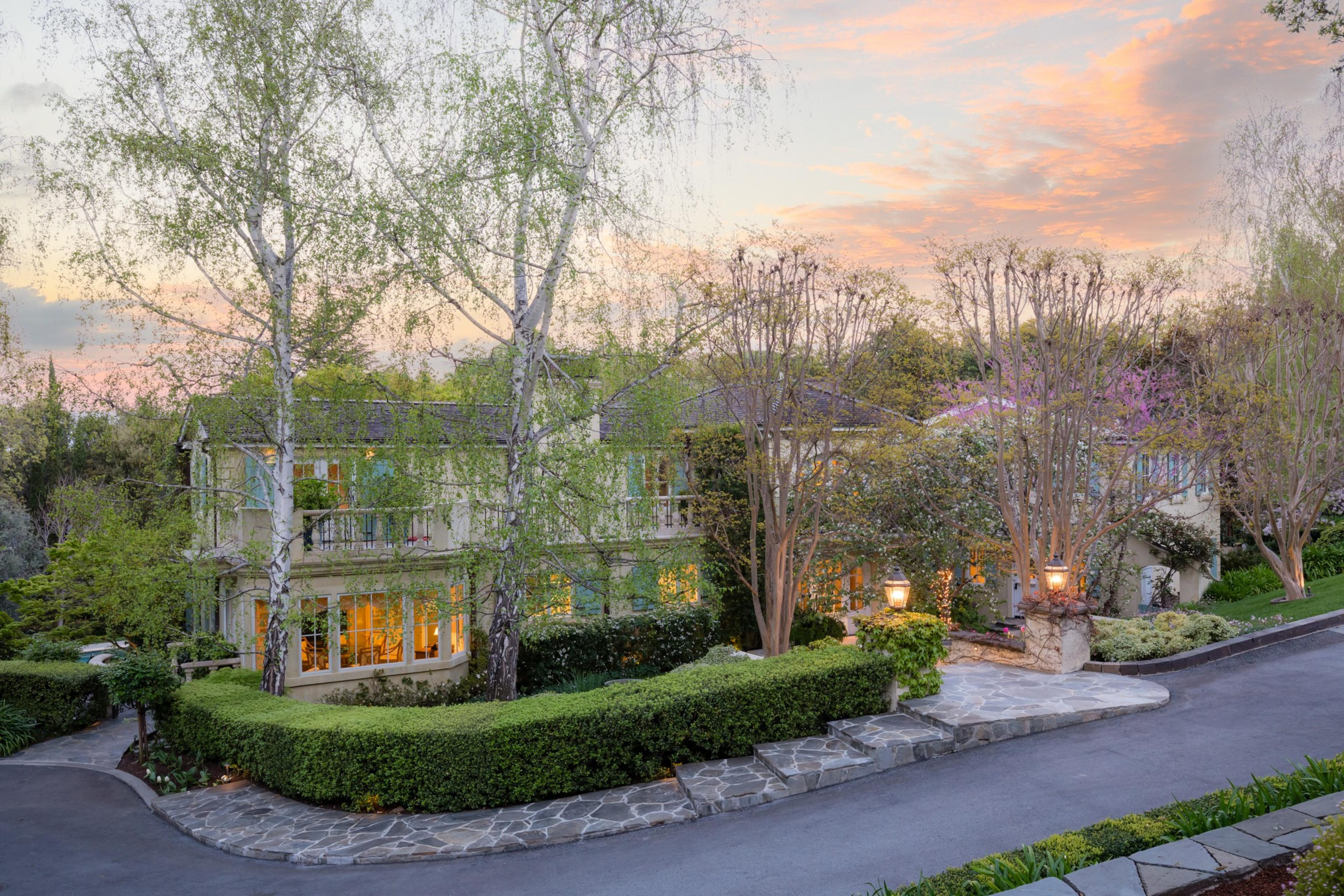 Elegant house with lights on, surrounded by trees, with a driveway at dusk under a pink-hued sky.