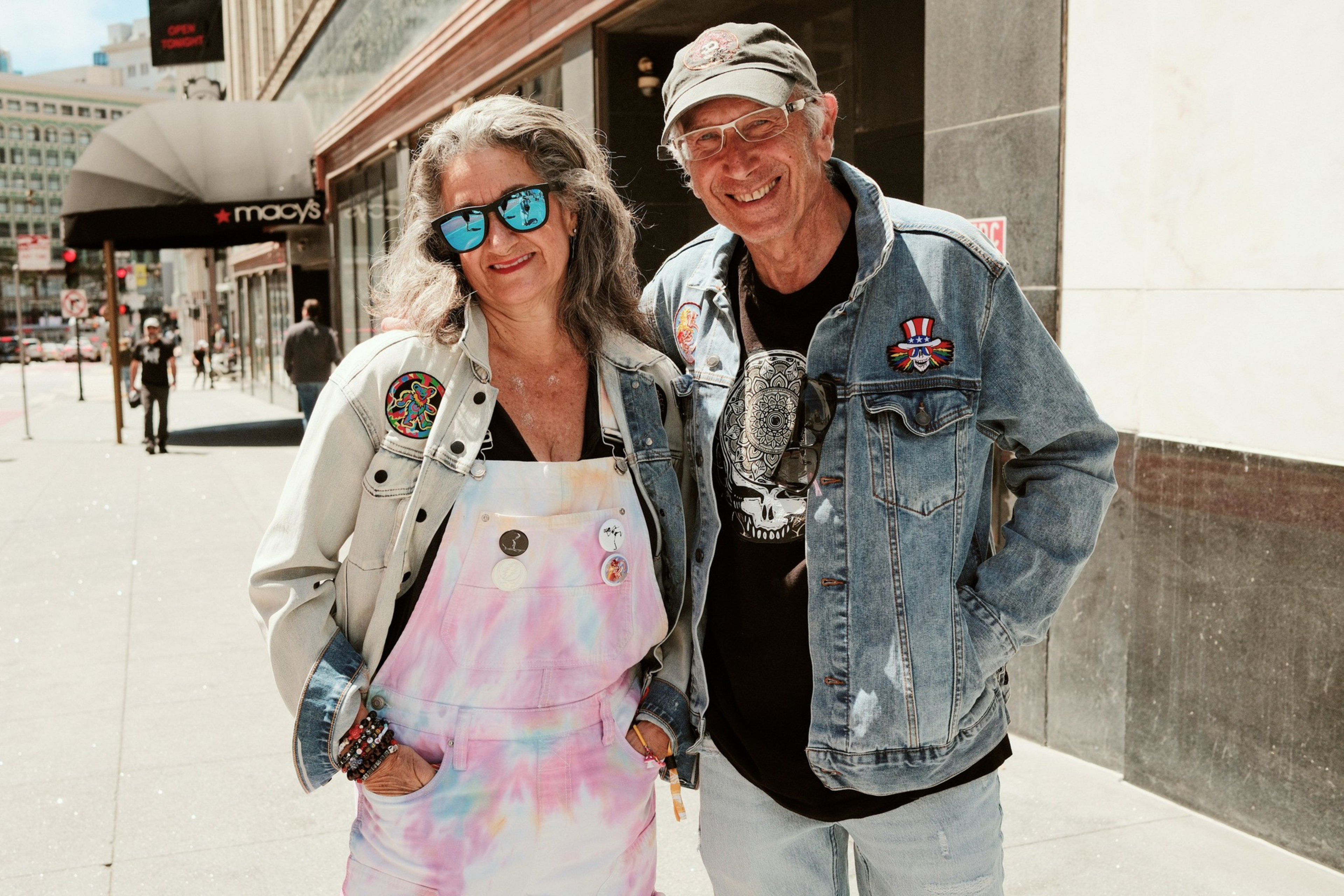 A smiling older couple dressed in casual, colorful clothing and denim jackets stands together on a sunny city sidewalk near a Macy’s store.