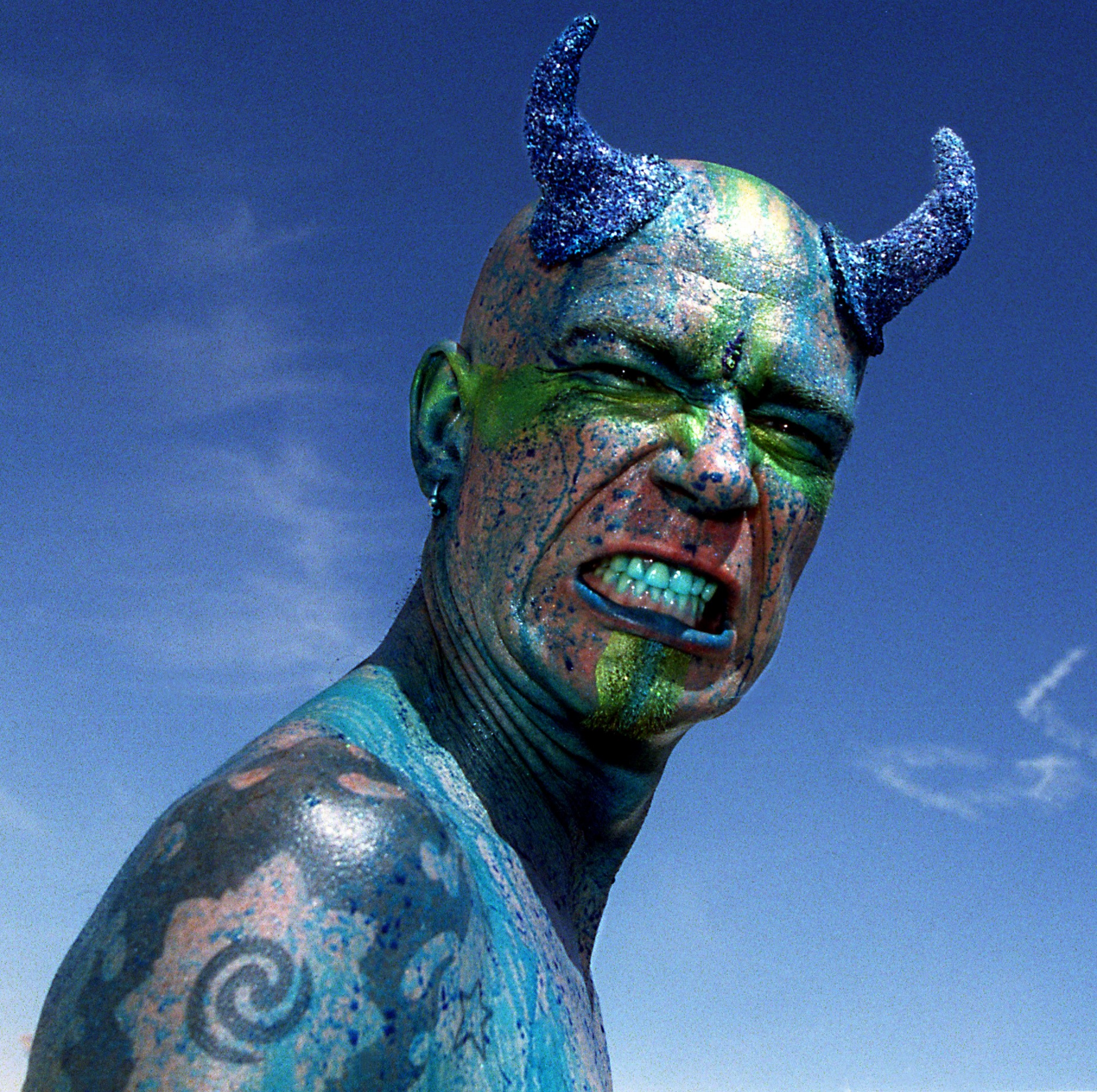 A person with glittery blue horns, blue and green face paint, and tattoos snarls aggressively against a clear blue sky.