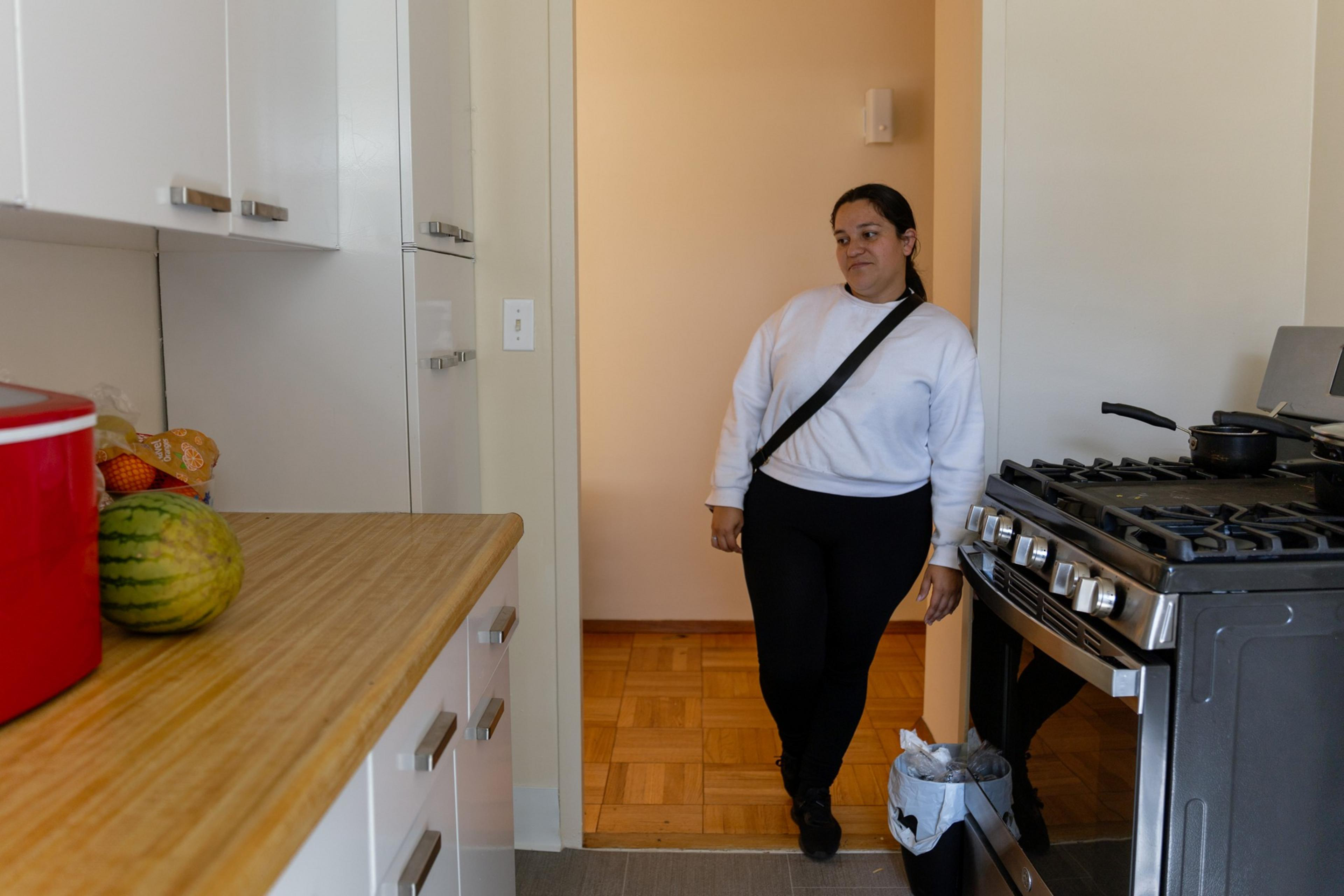 A woman in a white sweater and black pants stands in a kitchen doorway. The kitchen has white cabinets, a stove with pans, and a countertop with a watermelon.