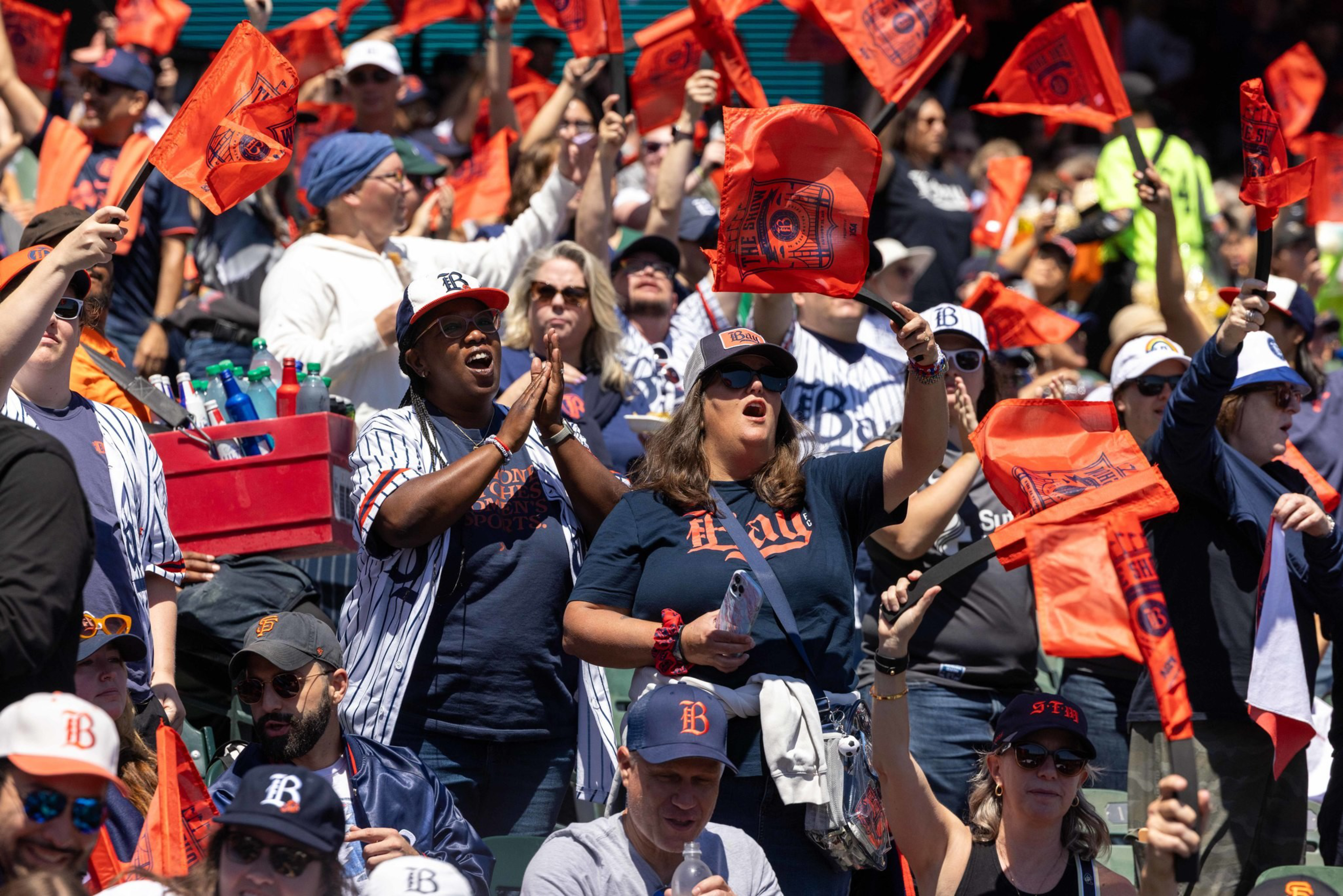A crowd of sports fans wearing caps and jerseys wave orange flags energetically, cheering and celebrating together in bright sunlight.