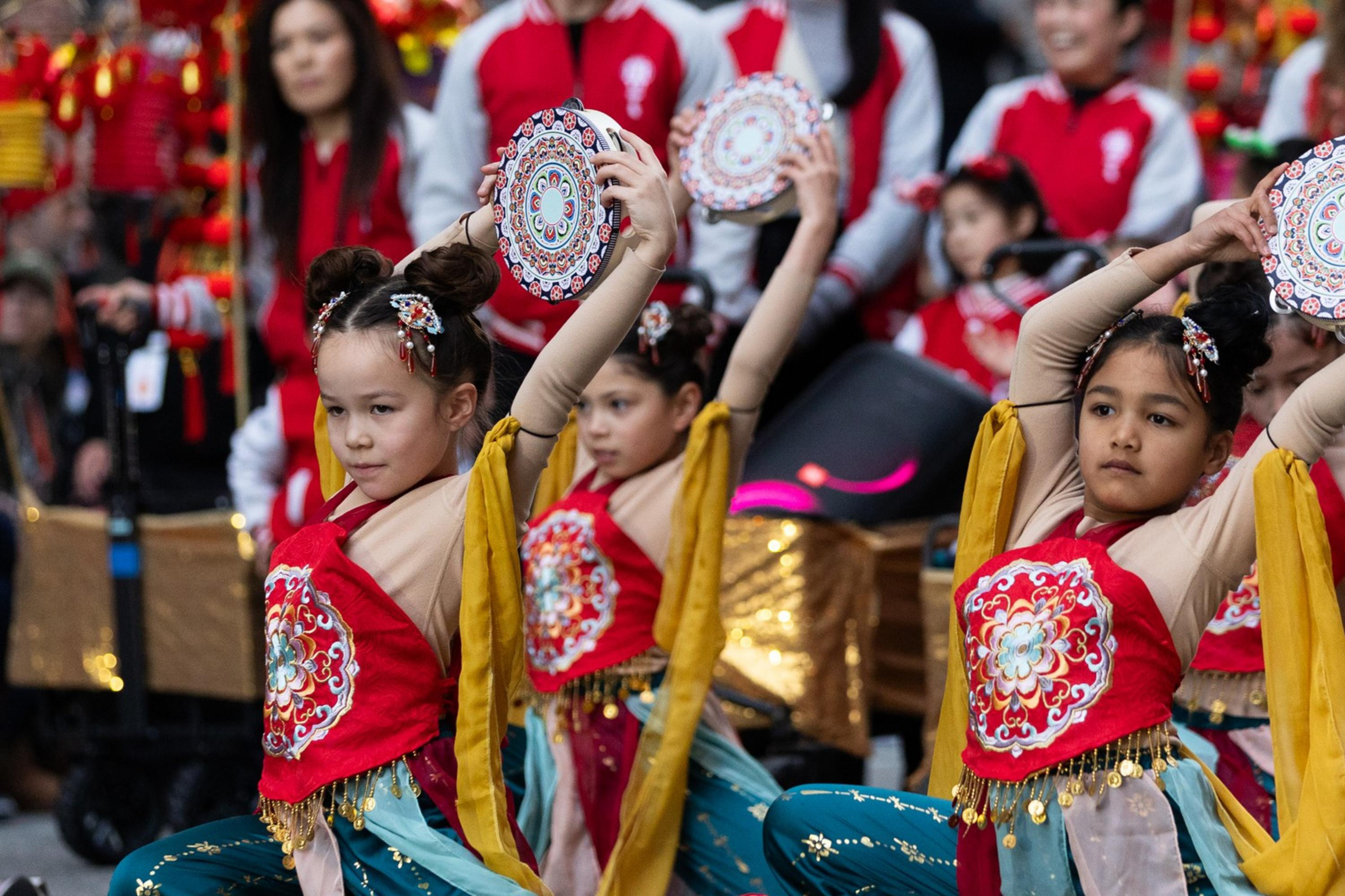 Children in colorful outfits perform a dance, holding decorated circular instruments aloft. They are kneeling on one knee with synchronized poses.