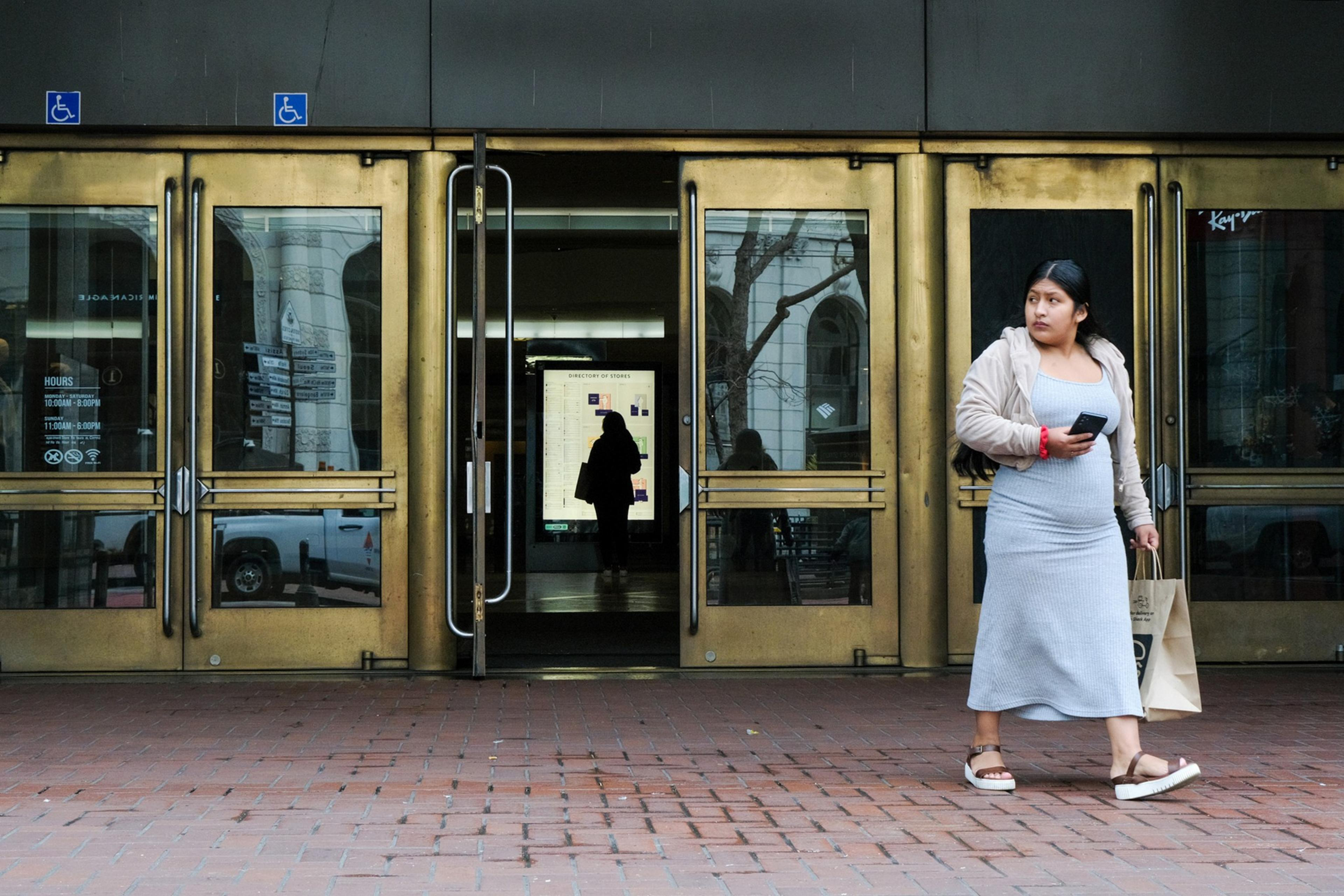 A woman walks past brass-colored building doors, holding a phone and a bag; reflection and interior visible.