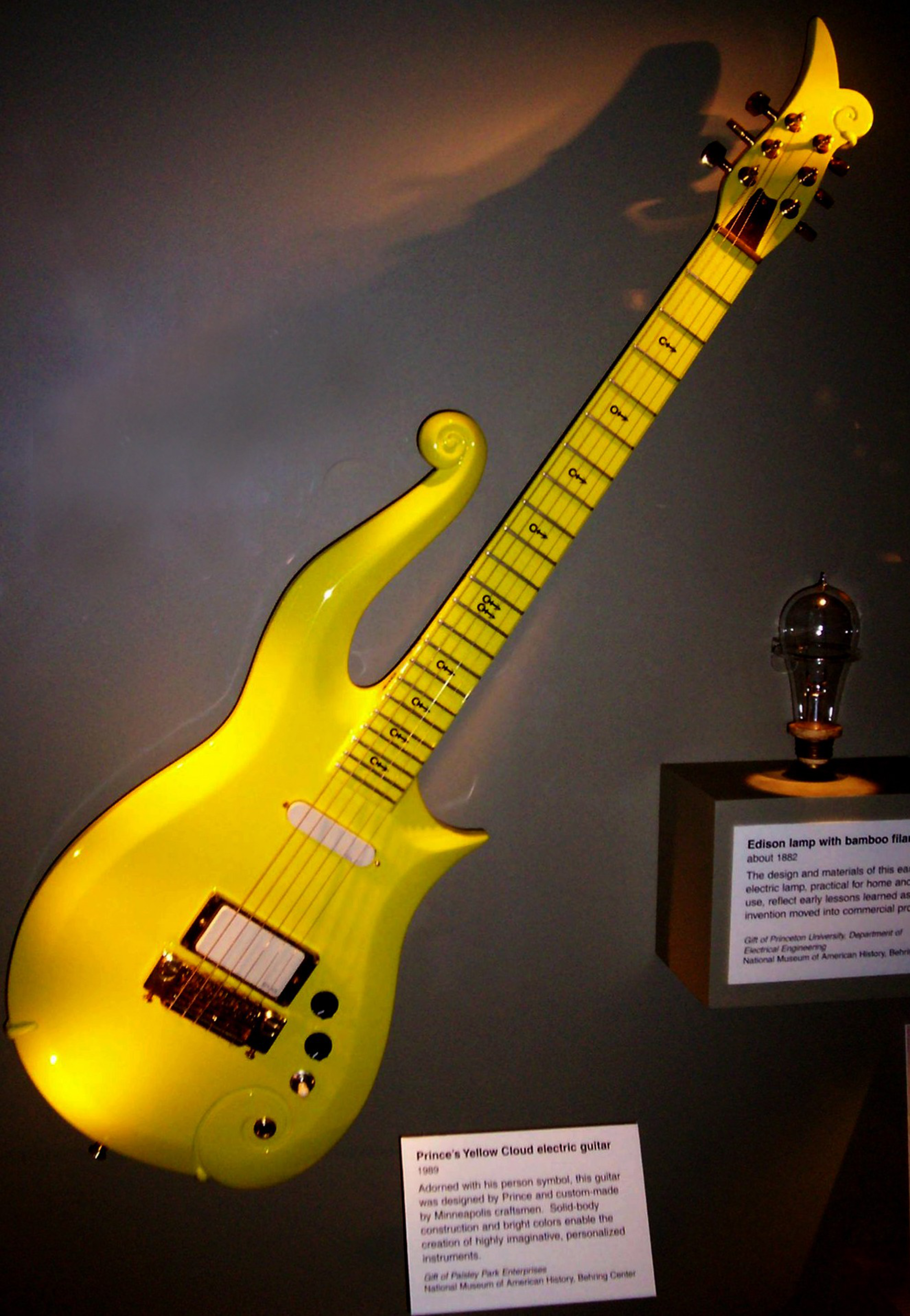 A bright yellow electric guitar with a unique, curvy body and scroll-shaped headstock, displayed in a museum with explanatory plaques nearby.