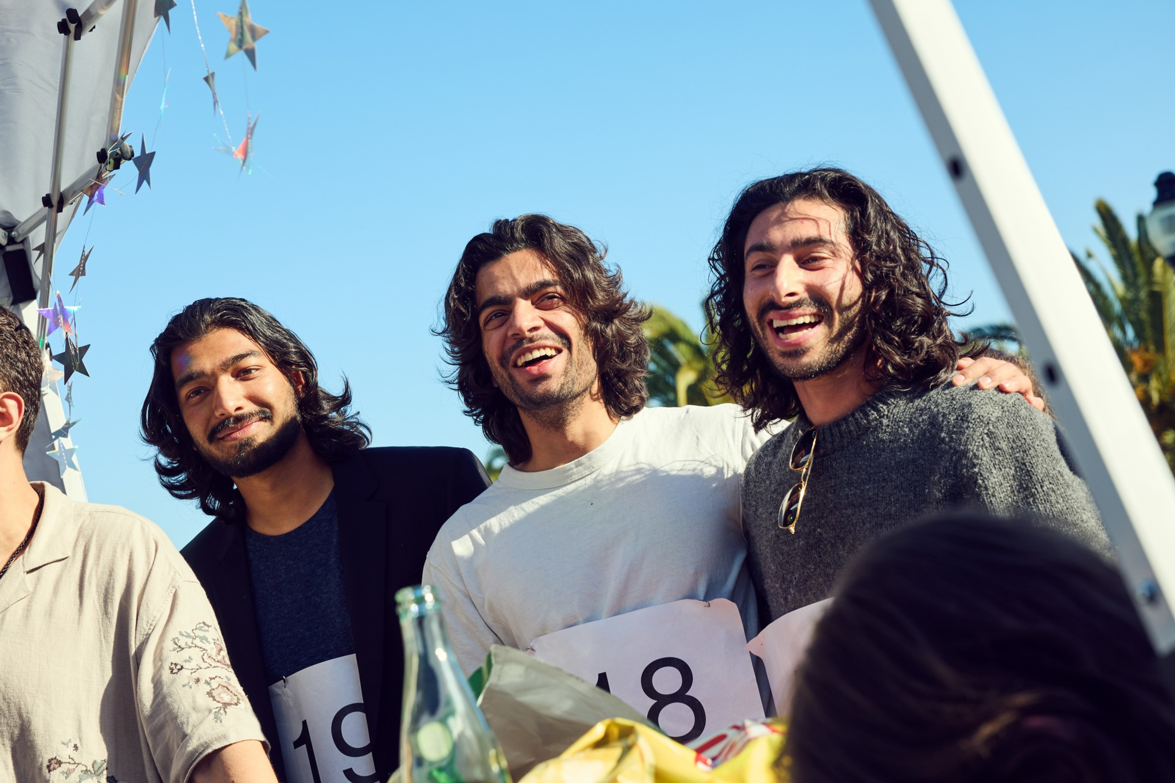 Three smiling men with long hair stand close together under a canopy decorated with star-shaped garlands against a clear blue sky. They hold number signs.