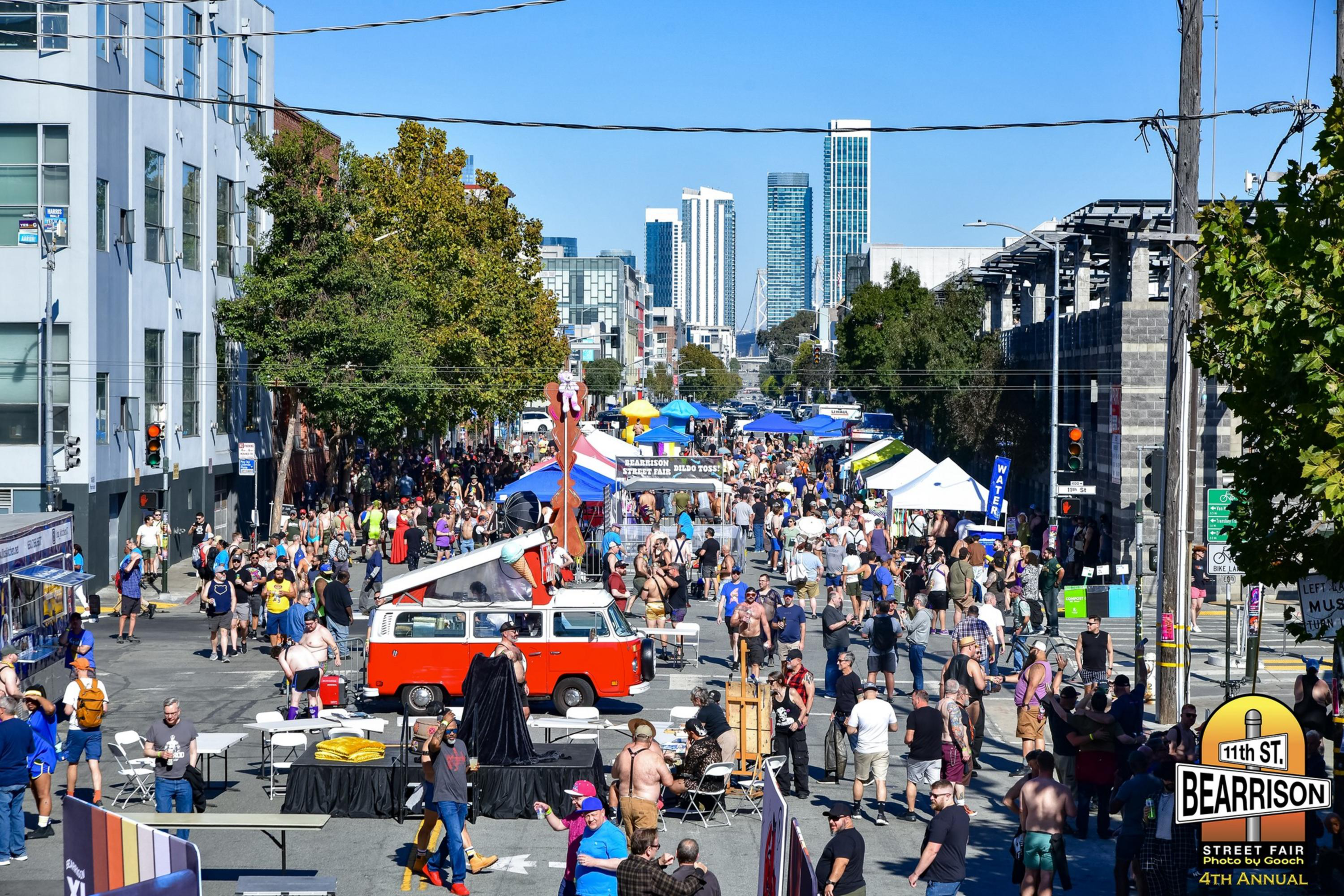 A crowded street fair with colorful tents, a red-and-white van, many people walking and socializing, trees, and tall buildings in the background under a clear blue sky.