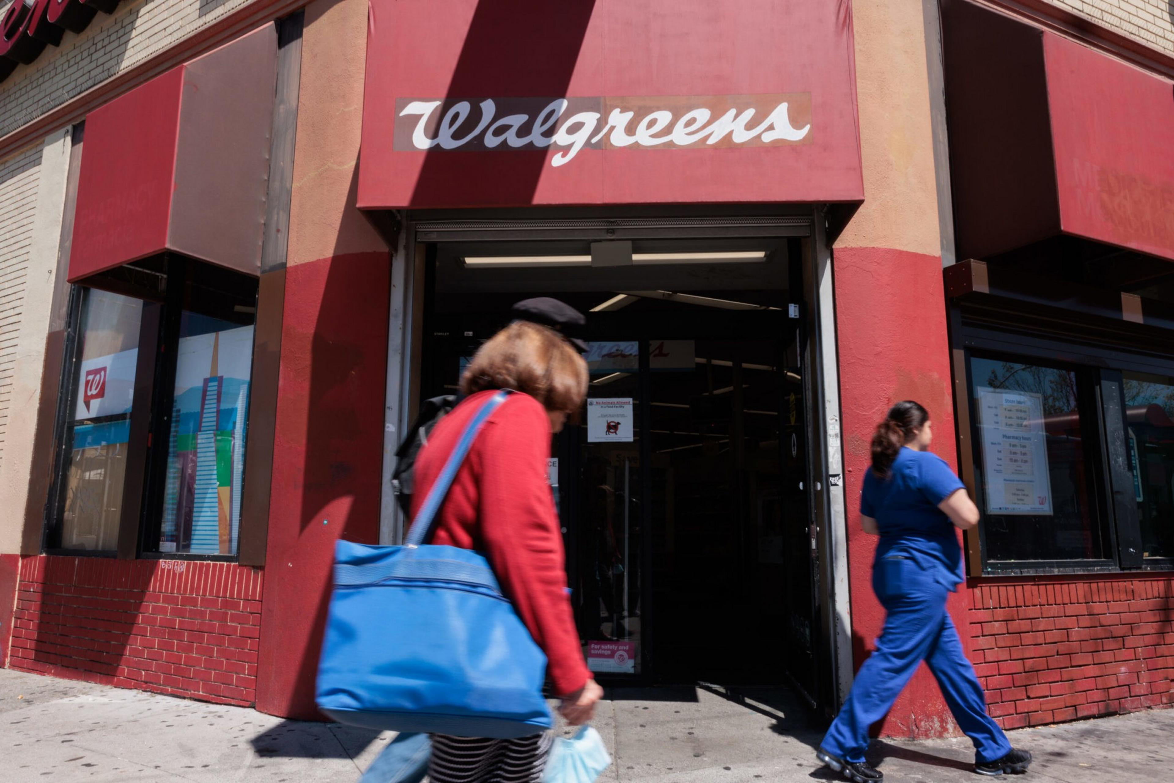 A Walgreens store entrance with a red facade. A woman in red with a blue bag walks past, and another woman in blue scrubs approaches the entrance.
