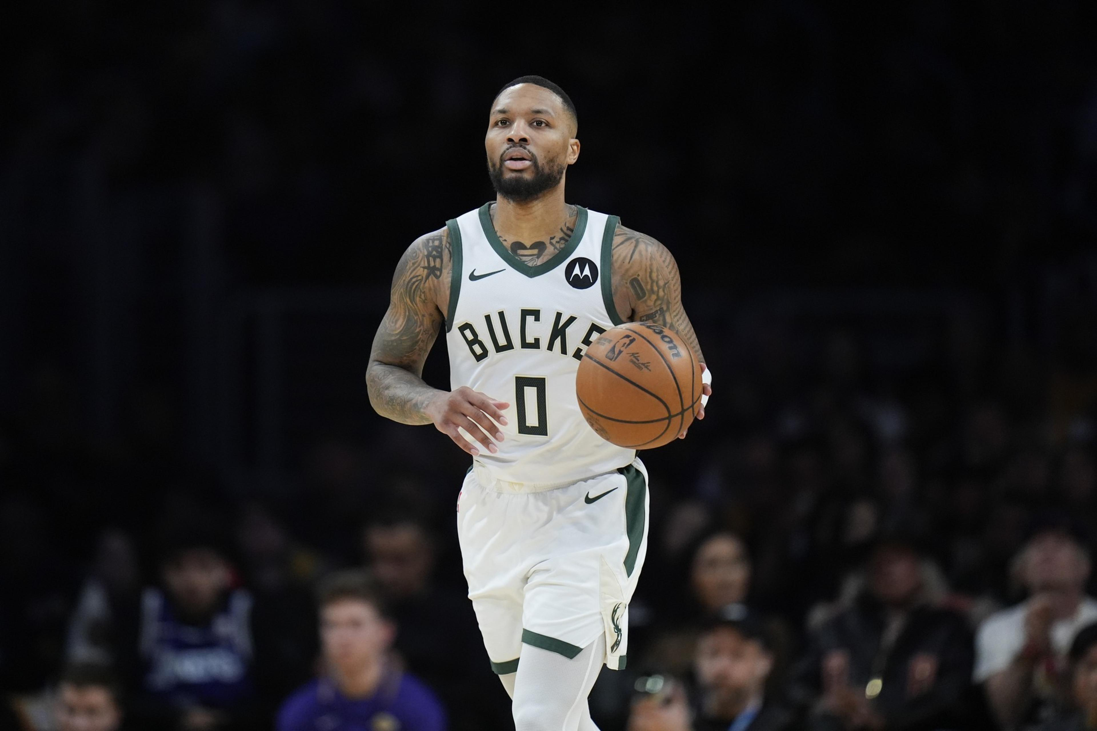 A basketball player wearing a Bucks jersey dribbles the ball on the court. He's focused and in motion, with a blurred crowd in the background.